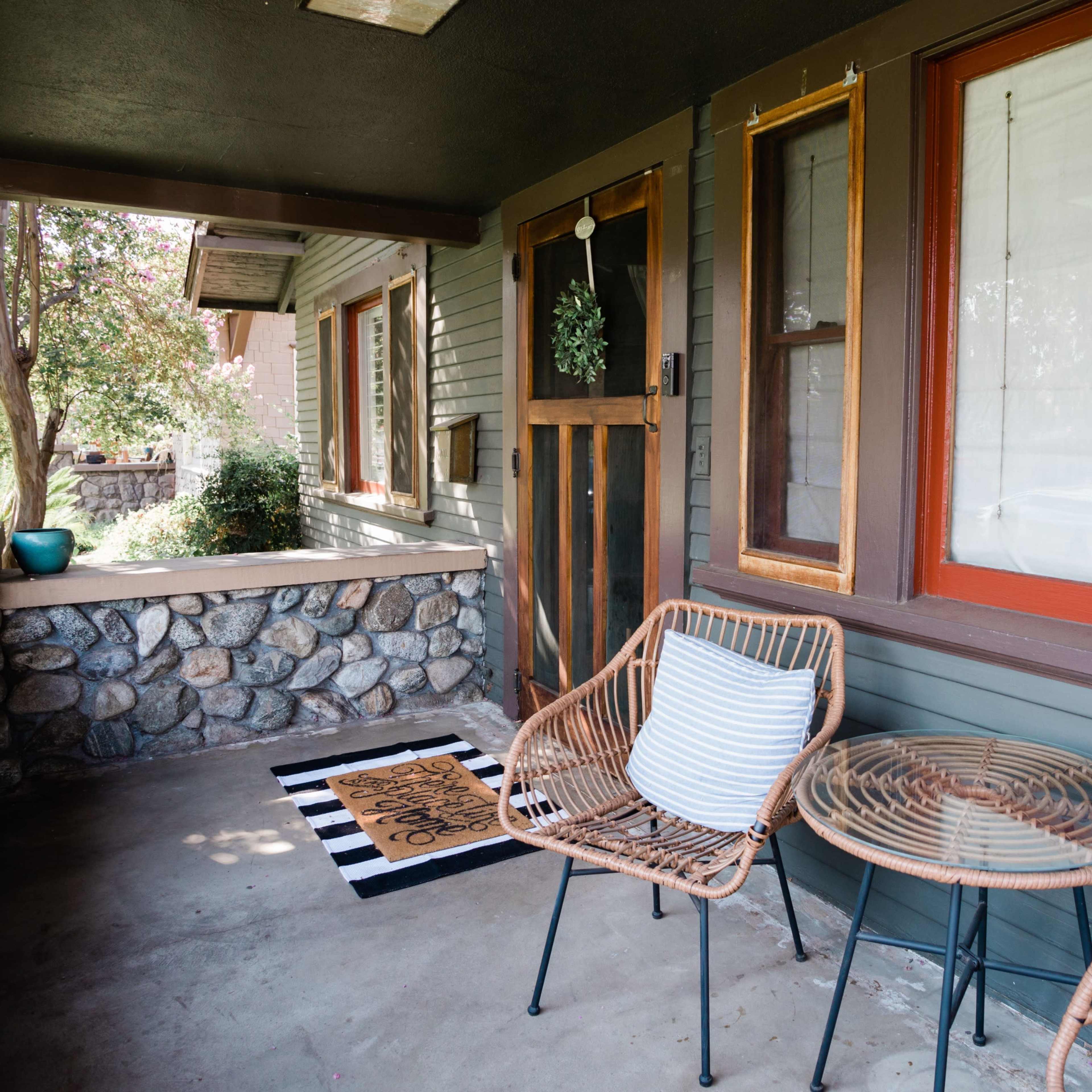 A porch with two wicker chairs and a small round table, set against a stone wall and featuring large windows with red frames.