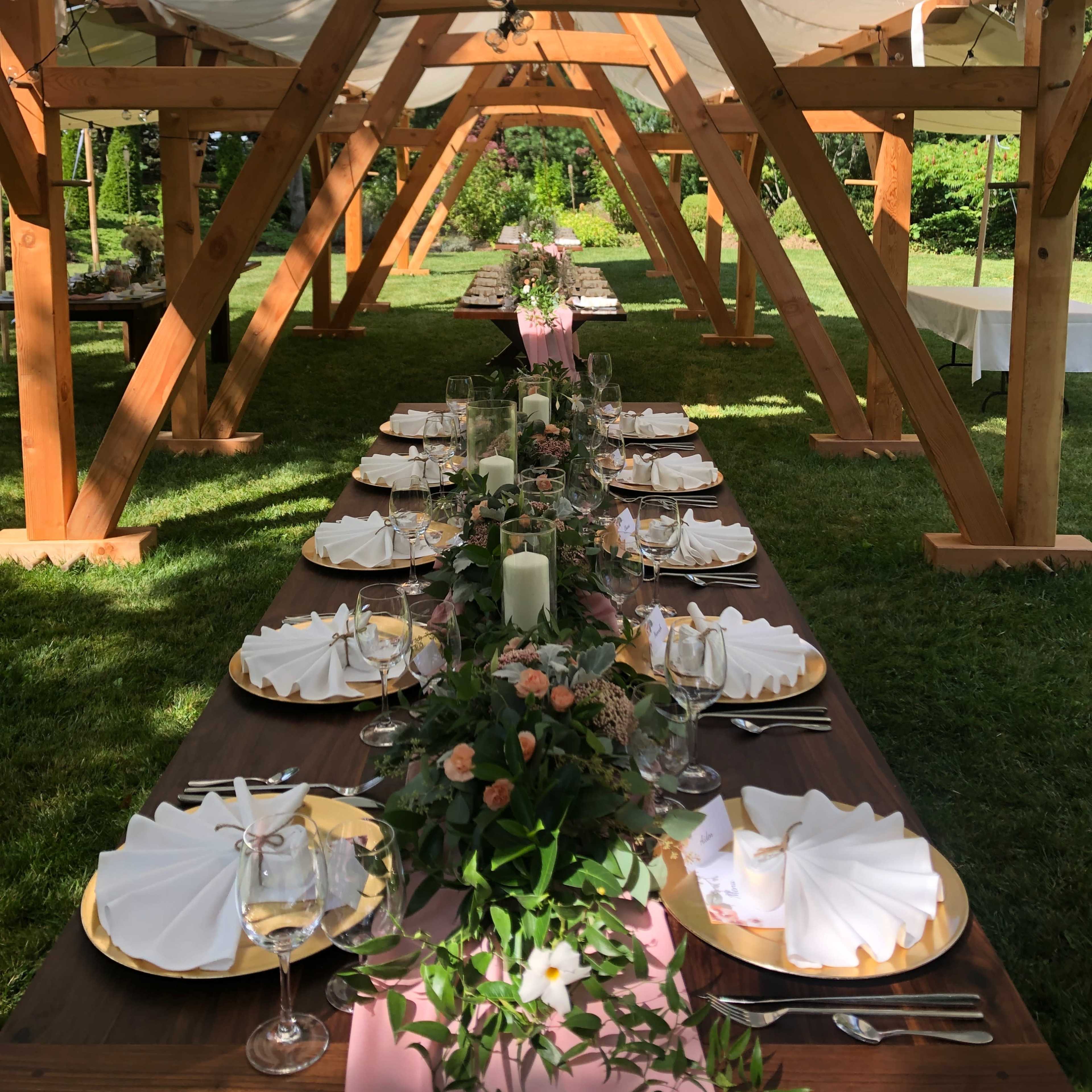 A long, elegantly set dining table under a wooden pavilion is surrounded by greenery.
