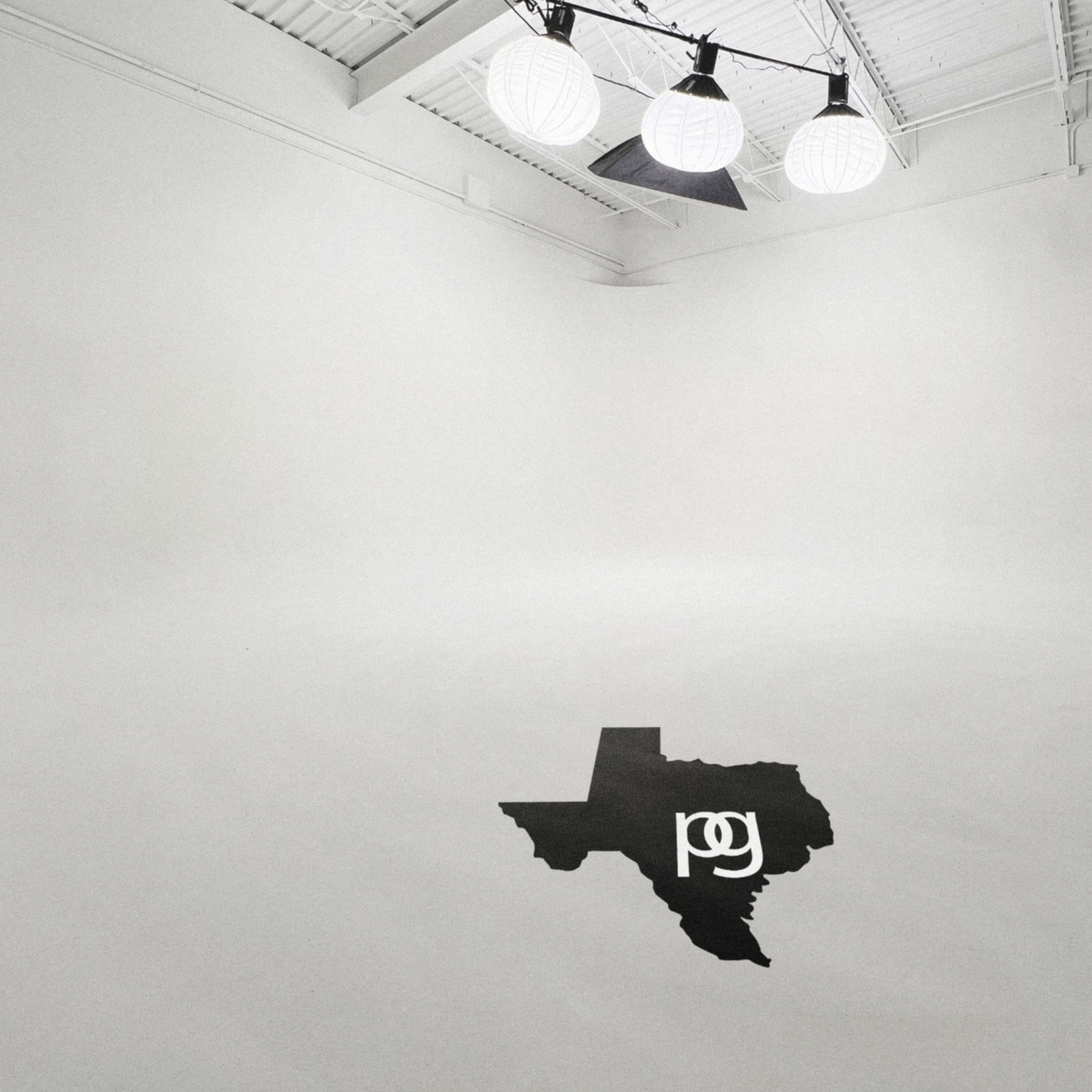 A spacious, empty studio with white walls and three hanging lights, featuring a black silhouette of Texas on the floor.