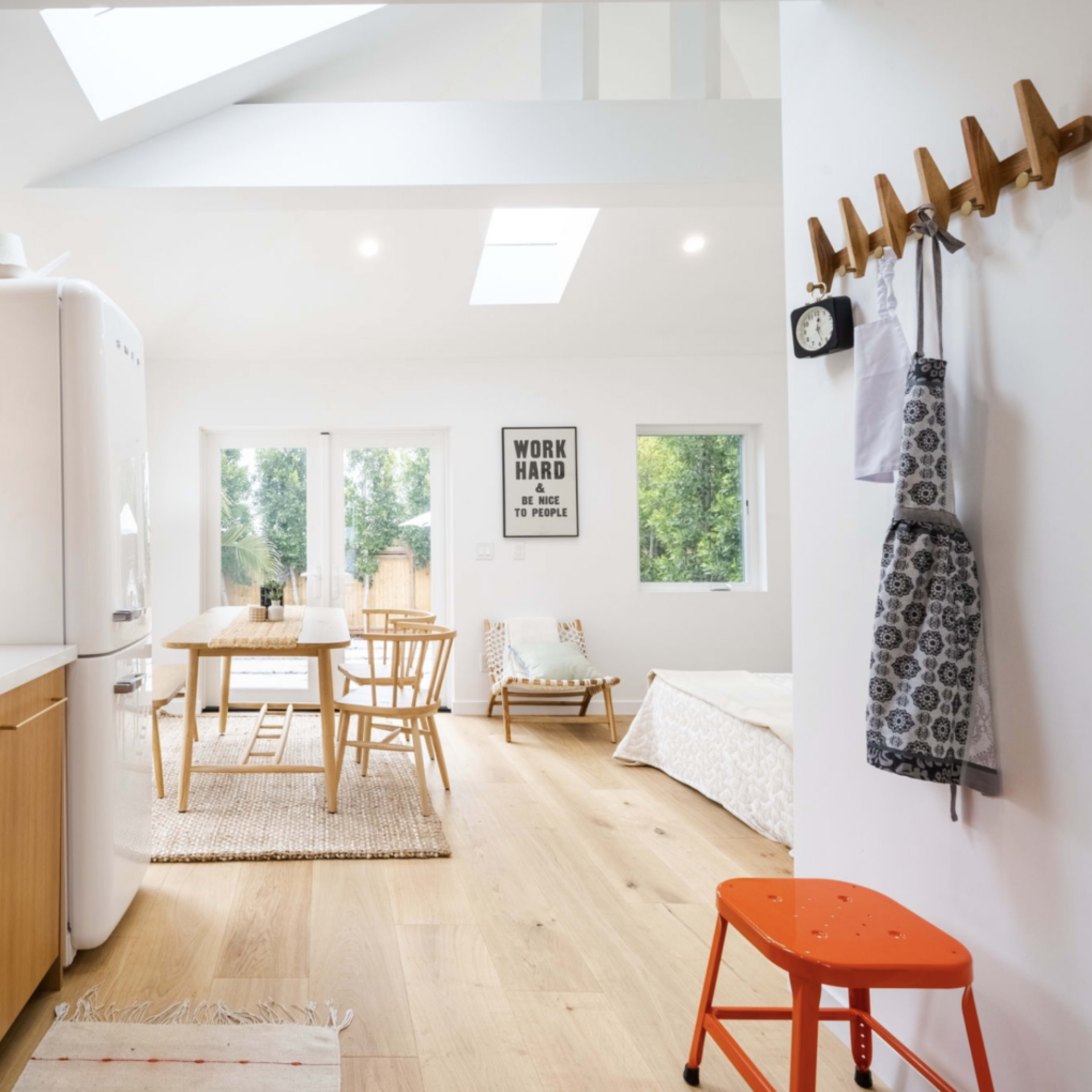 A bright, open-concept kitchen and dining area features wooden shelves, a white refrigerator, a dining table with chairs, and access to a cozy living space.