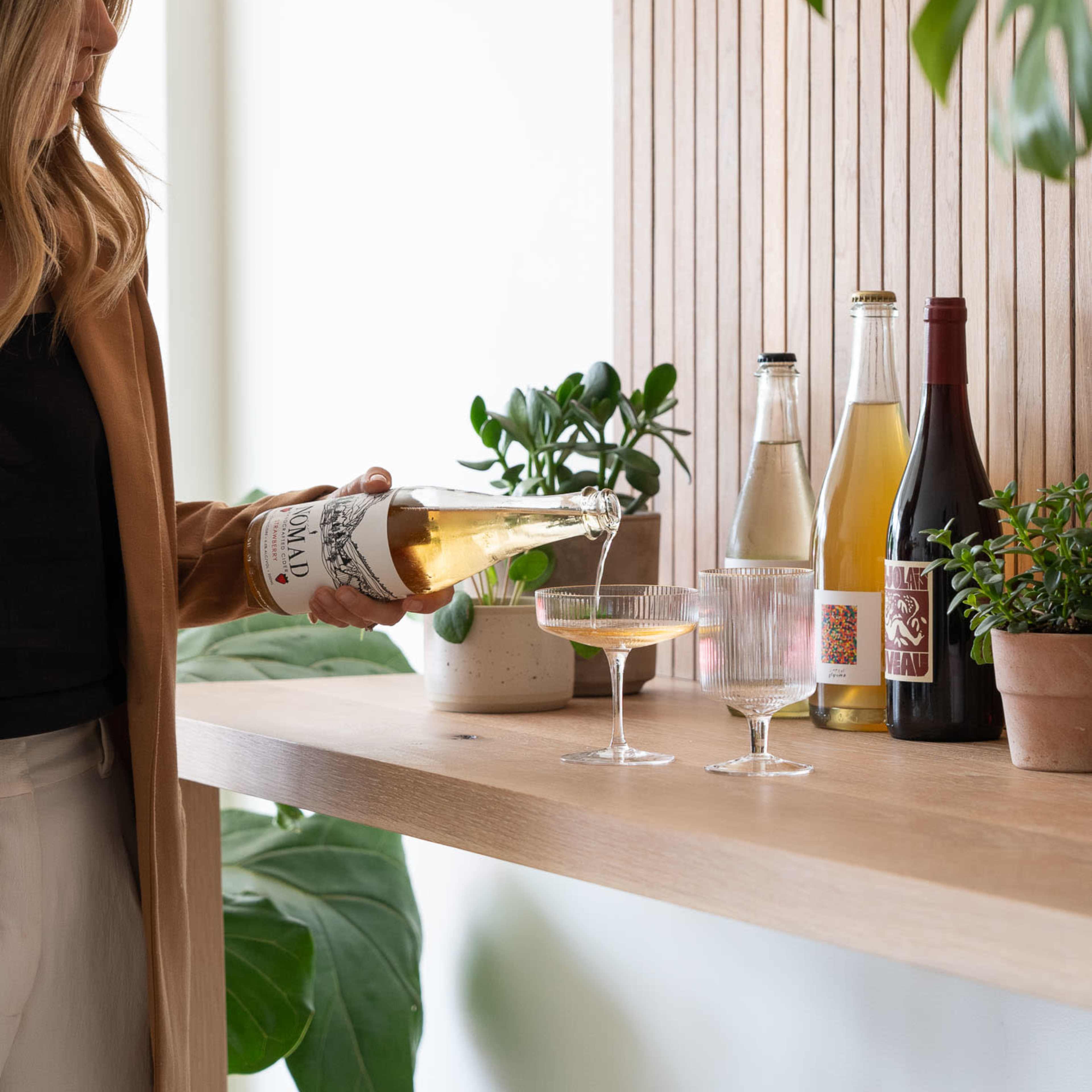 A woman pours a drink from a bottle into a glass on a wooden shelf adorned with various beverages and plants.