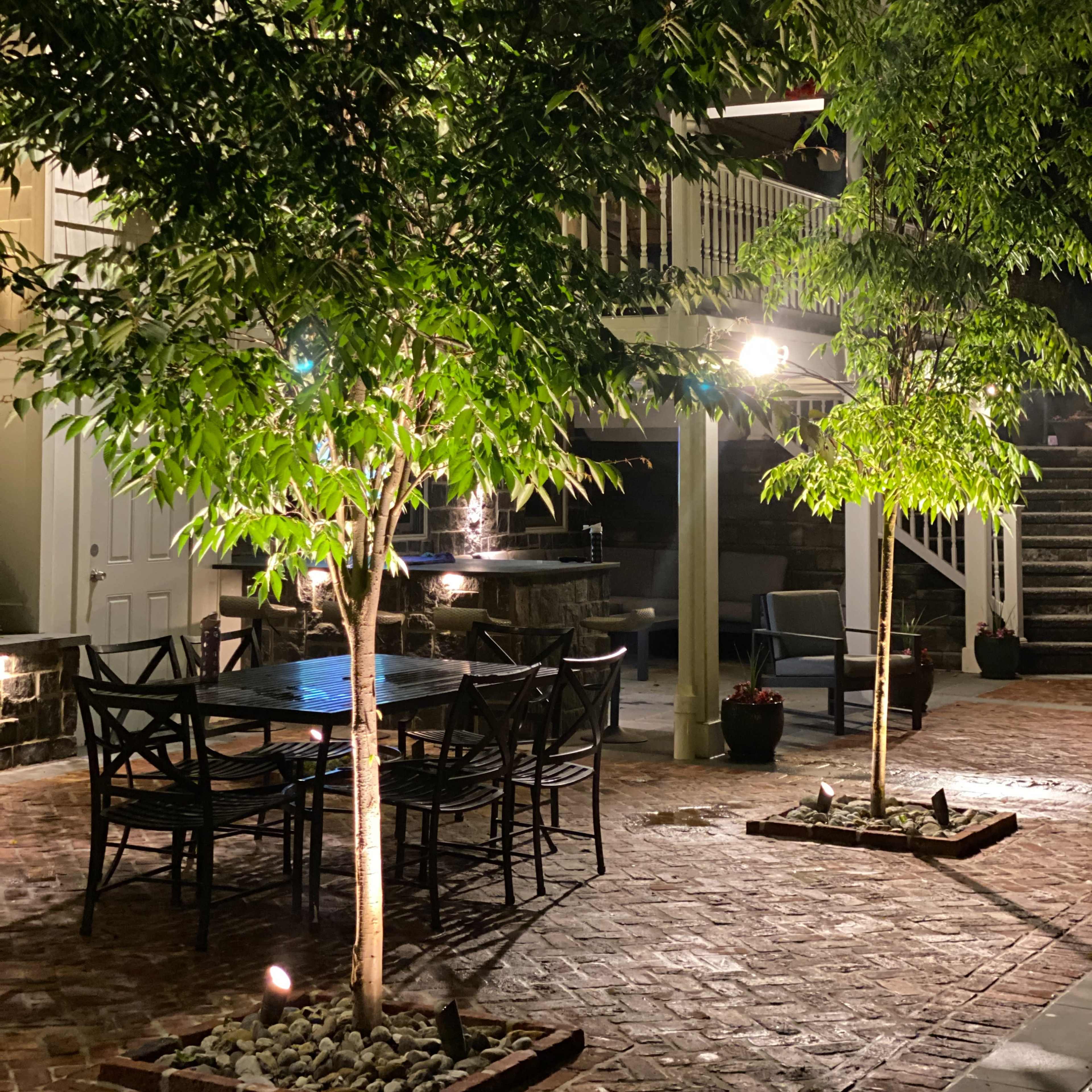 A well-lit patio with two trees surrounded by stone borders and a dining set at night.