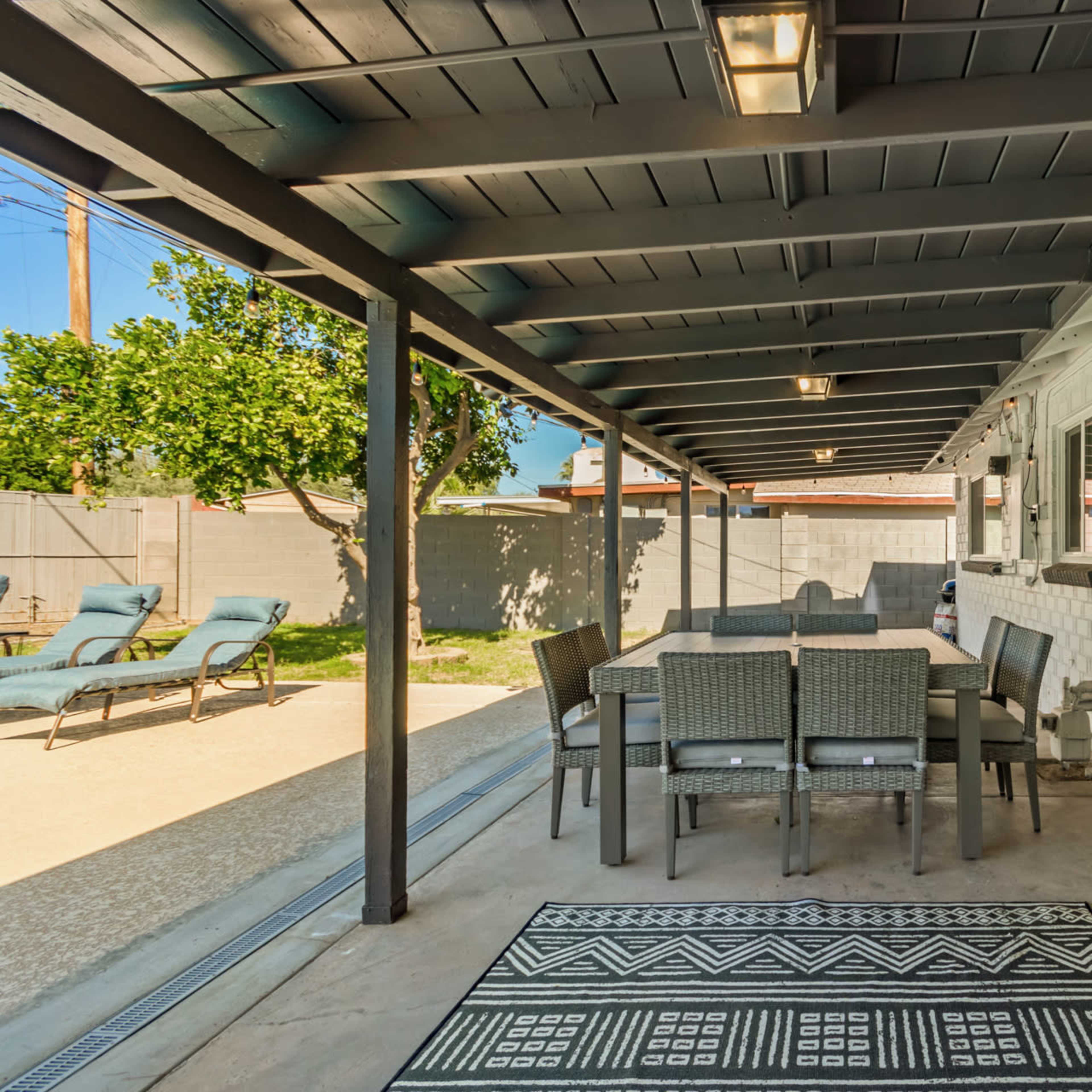 A covered patio with a dining table and chairs, a patterned rug, and lounge chairs in a yard with green grass.
