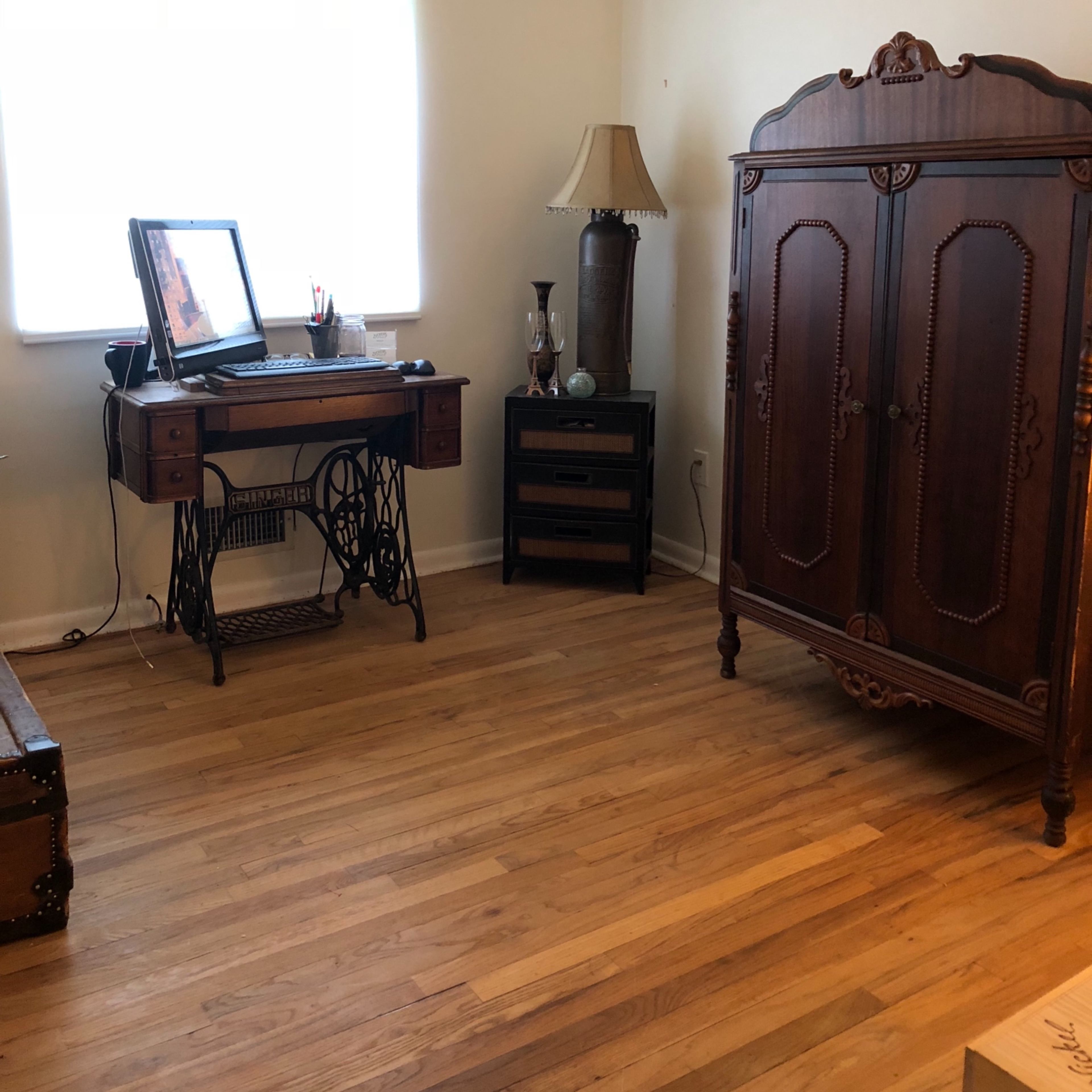 The room features a desk with a computer, a lamp, a wooden wardrobe, and a window allowing natural light to enter.