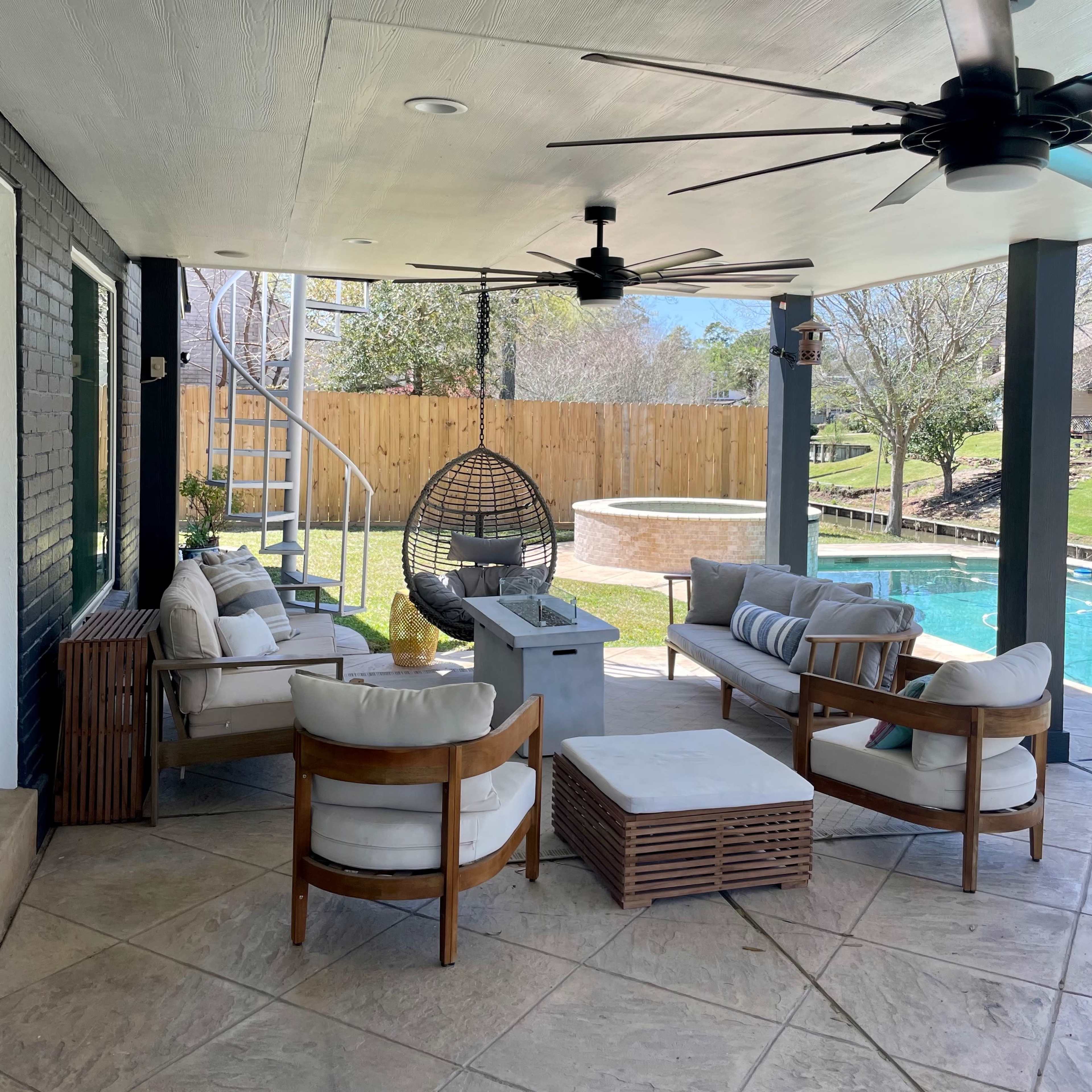 A covered patio area with comfortable seating arrangements, a hanging chair, ceiling fans, and a view of a swimming pool and grassy yard.