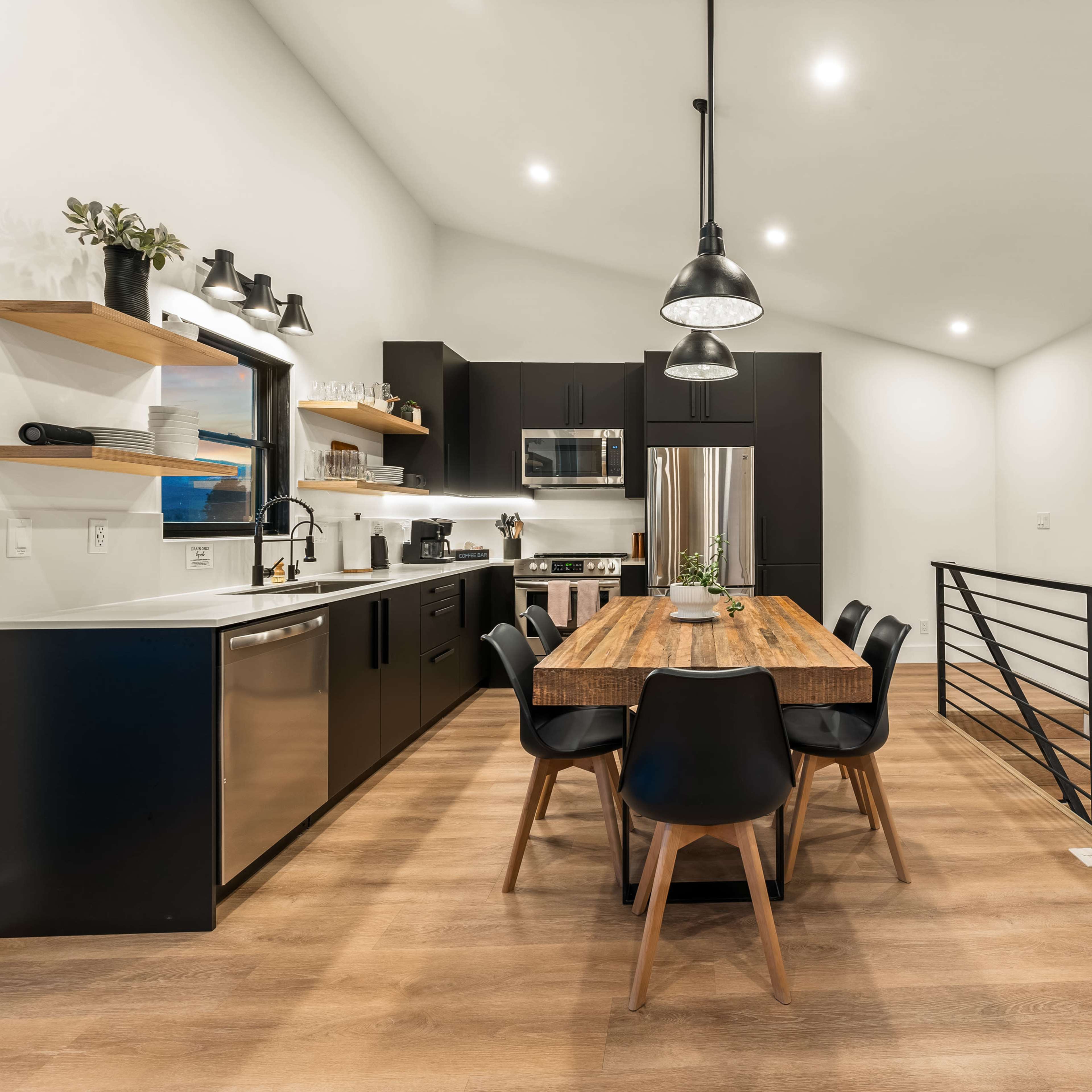 A modern kitchen features a black cabinetry design, a wooden dining table surrounded by black chairs, and large windows providing ample light.