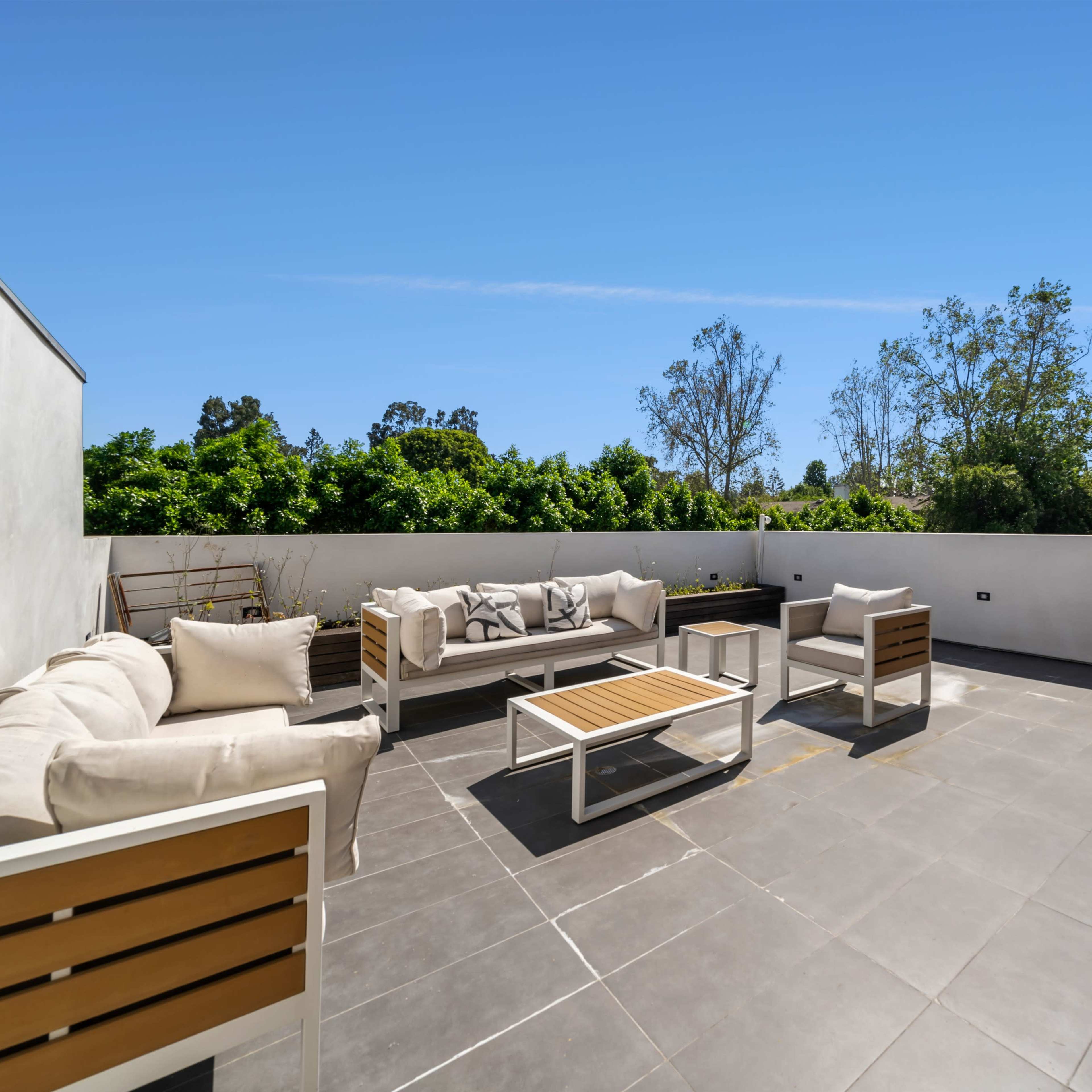 A modern rooftop terrace features patio furniture arranged on tiled flooring, surrounded by greenery and a clear blue sky.