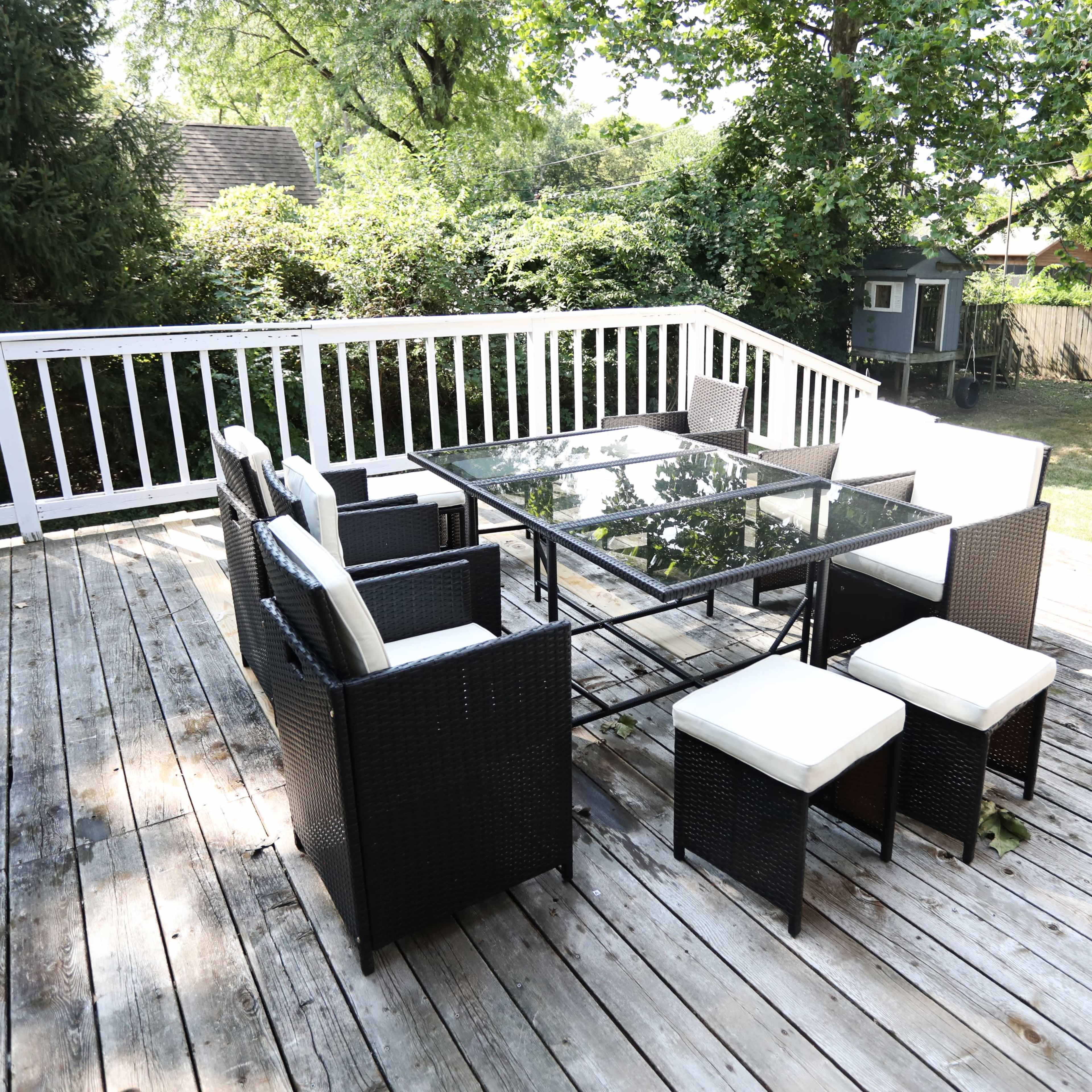 A black wicker patio set with a glass table and white cushions is arranged on a wooden deck surrounded by greenery.