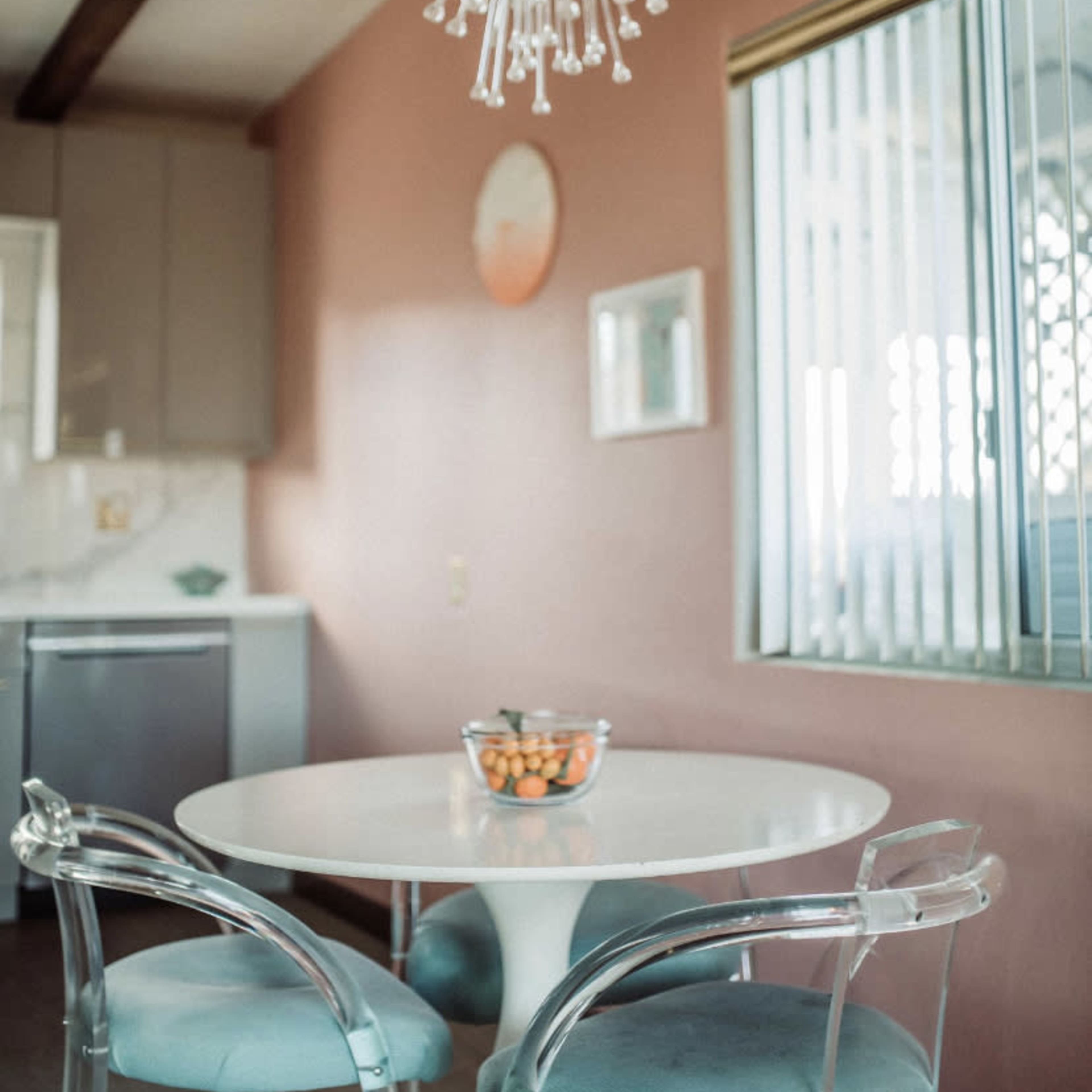 A modern kitchen features a round white table with transparent chairs and a bowl of oranges, illuminated by a distinctive chandelier.