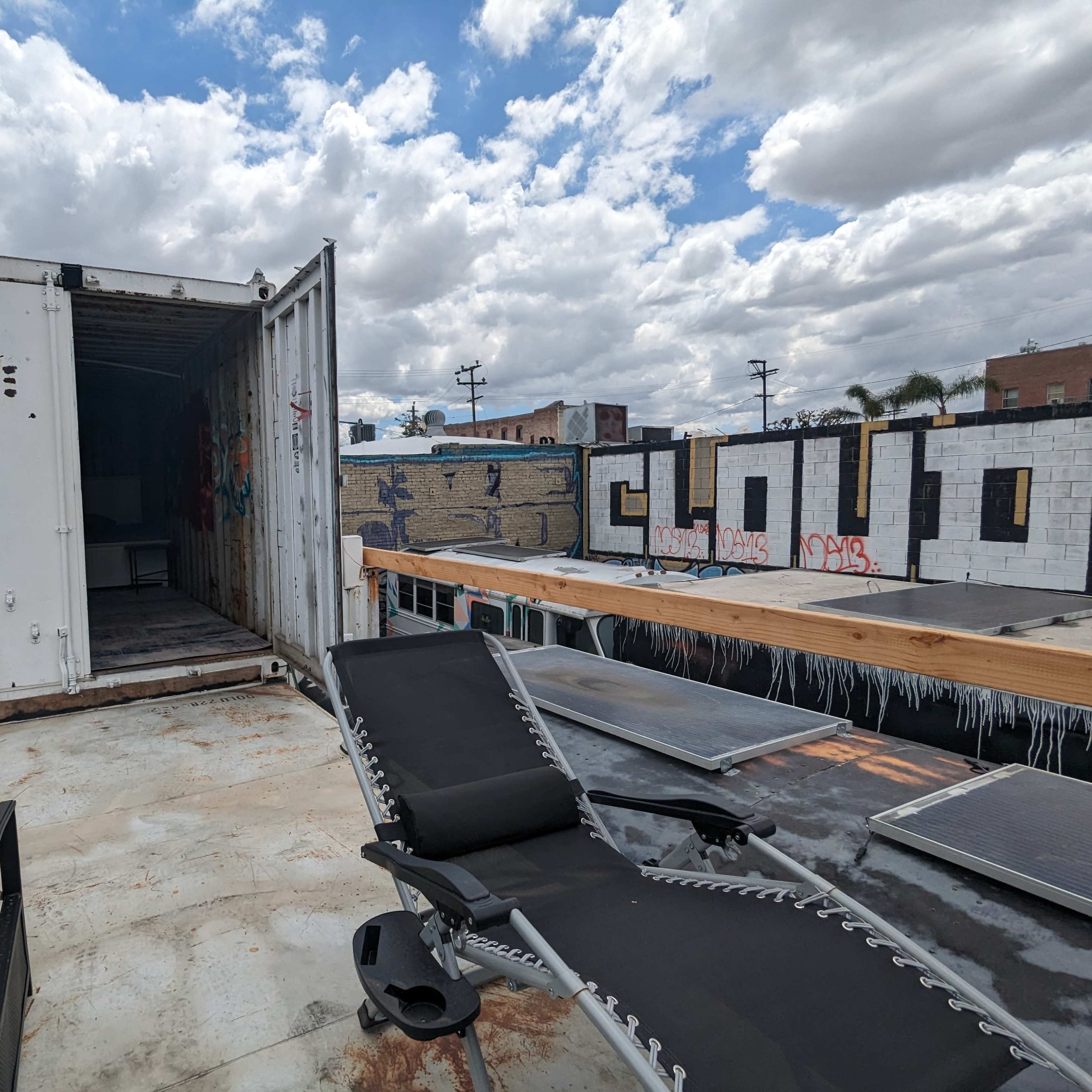 A rooftop area with a shipping container partially open, some chairs, and solar panels, set against a backdrop of graffiti-covered walls and cloudy skies.