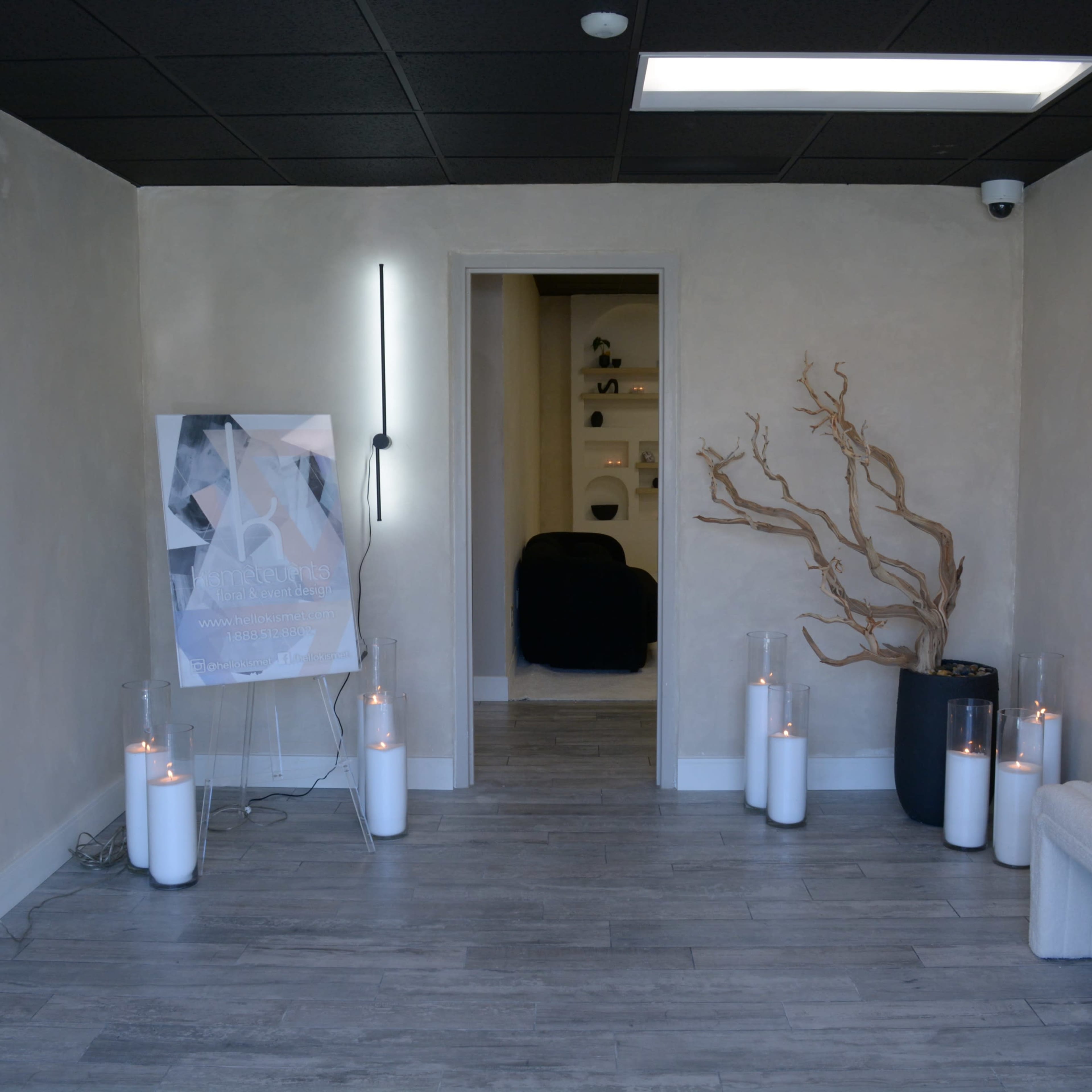 A minimalist interior features a bench, decorative branches, and candles in glass holders, with a poster and an open doorway leading to another room.