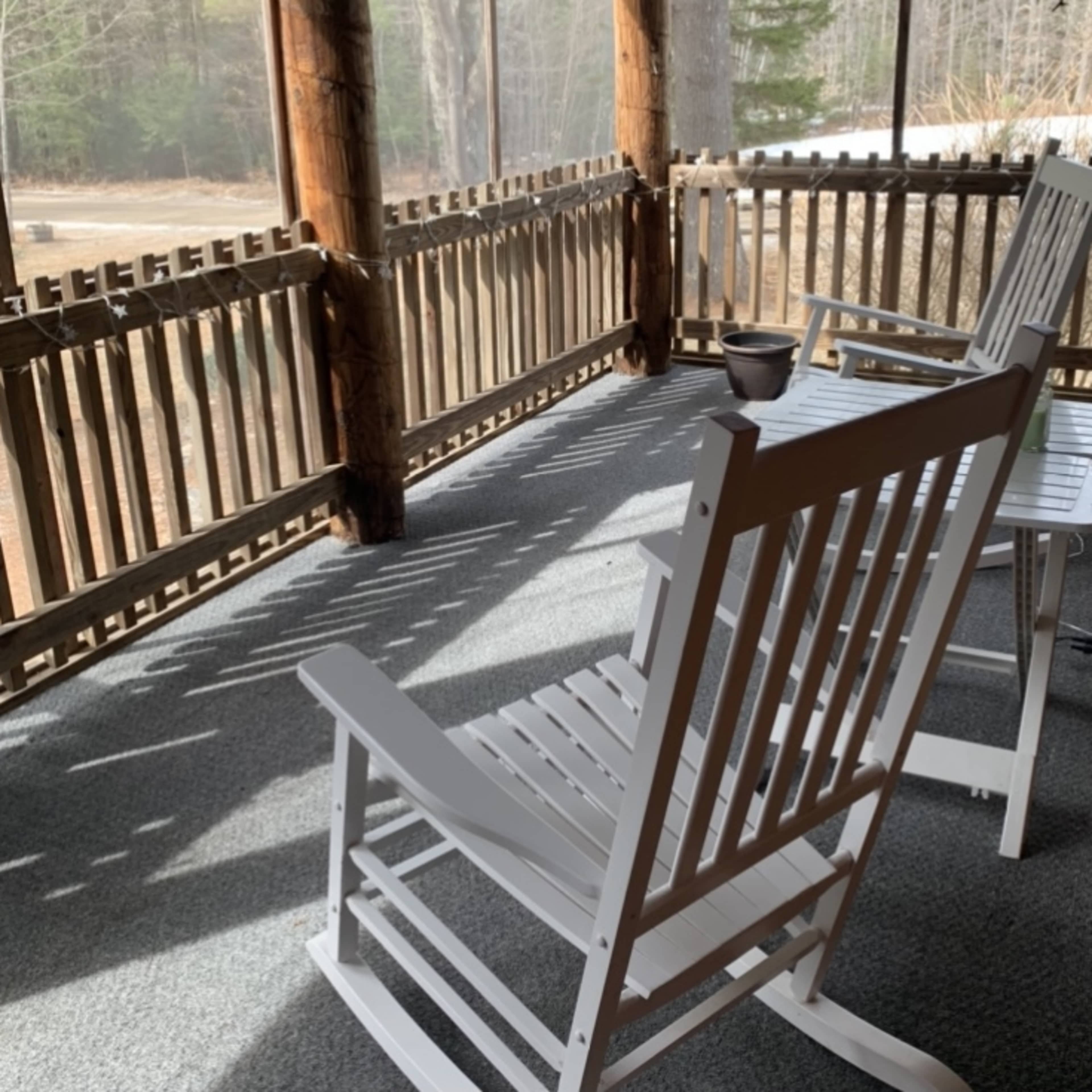 Two white rocking chairs are positioned on a screened porch with a view of a wooded area.
