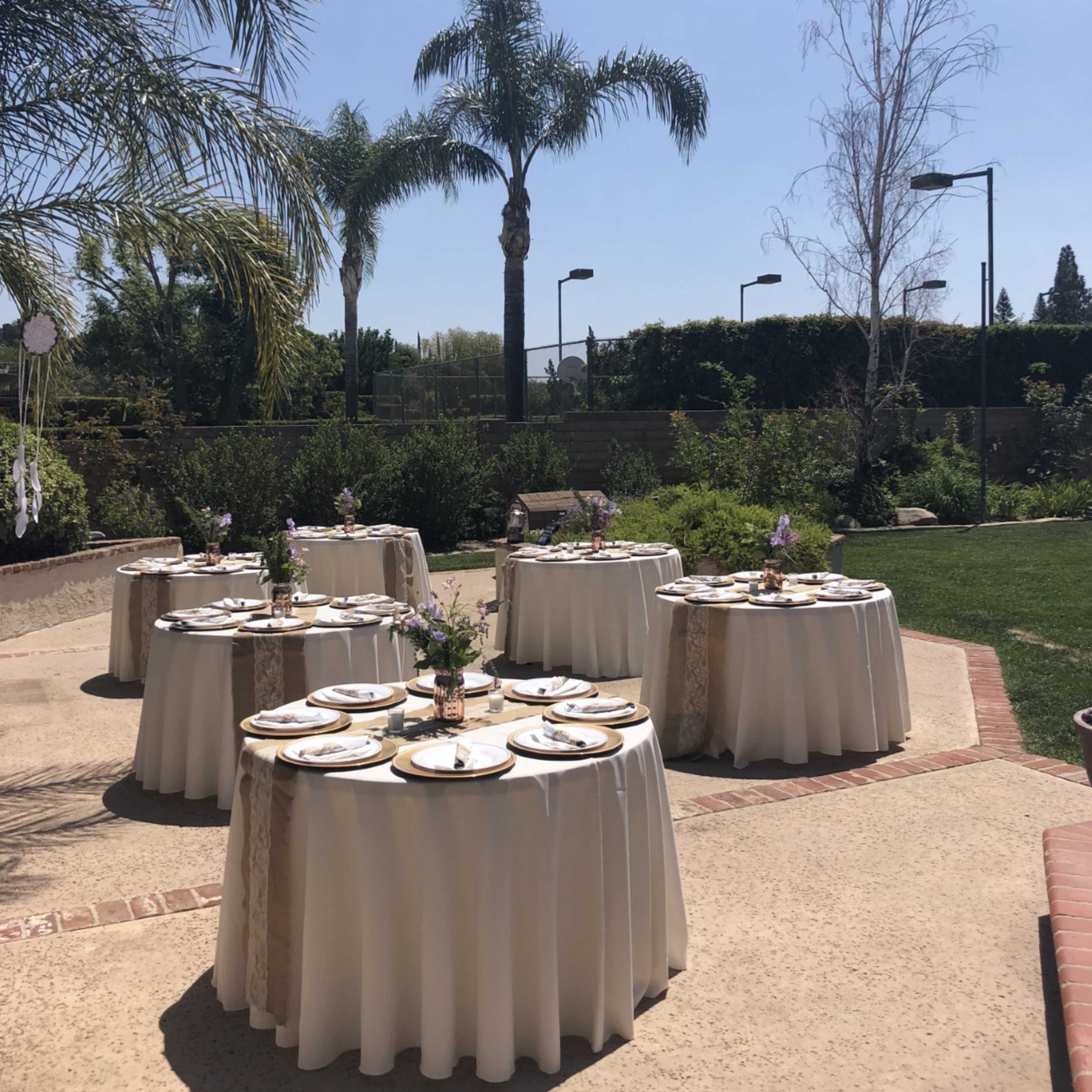 Several round tables with white tablecloths set up outdoors, surrounded by greenery and palm trees under a clear blue sky.