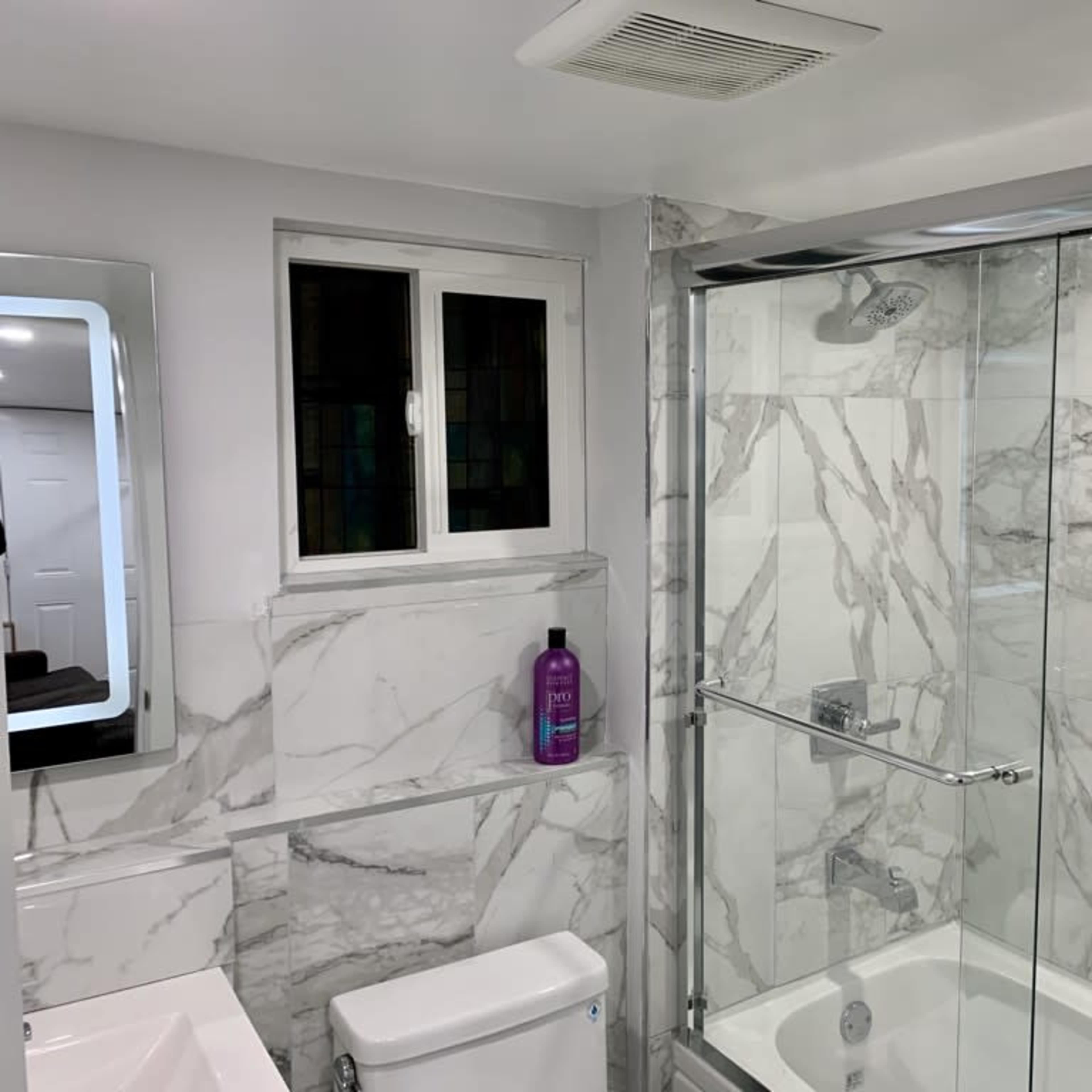 A modern bathroom featuring a glass shower enclosure, marble-patterned walls, a toilet, and a large mirror above the sink.