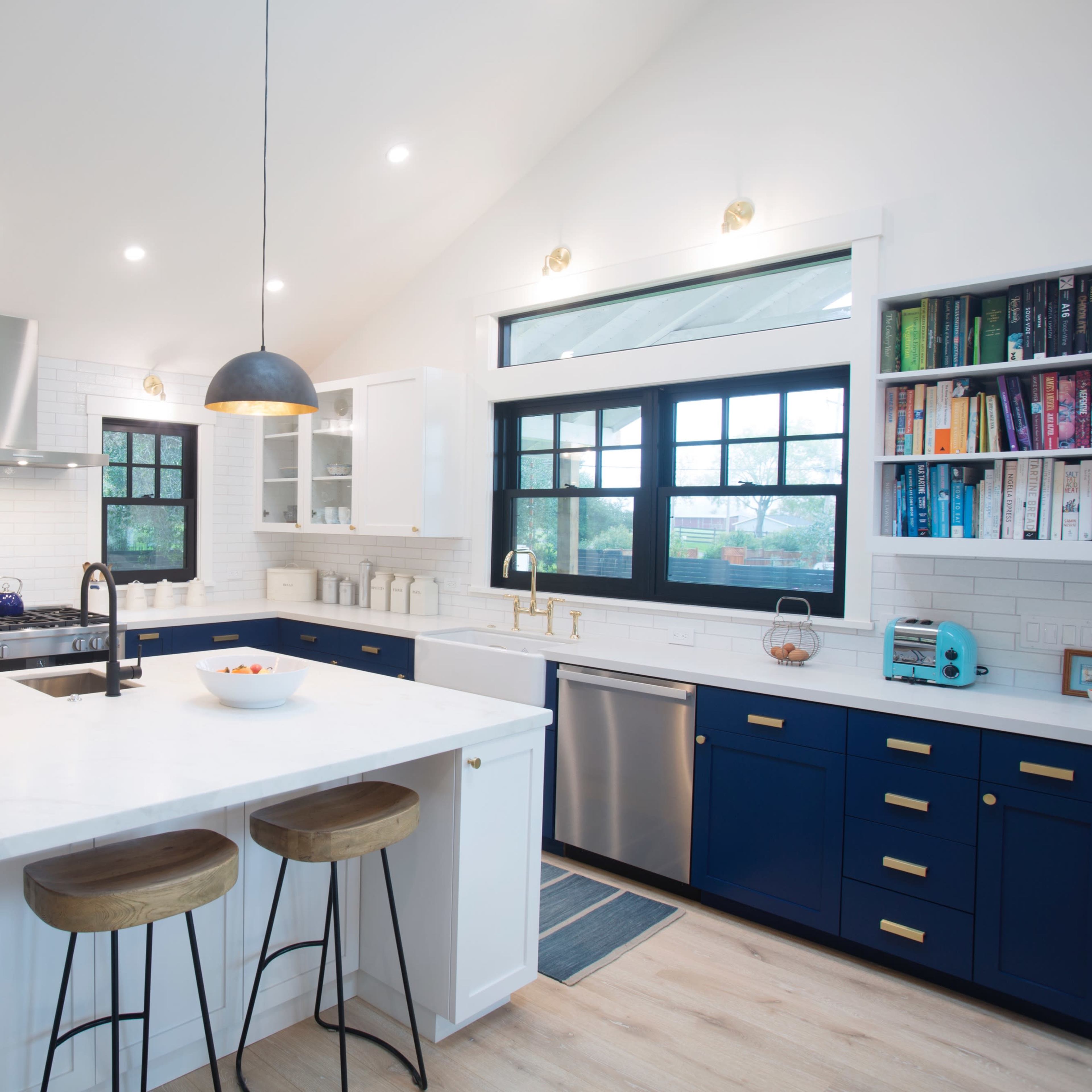 A modern kitchen features a central island with bar stools, blue cabinetry, stainless steel appliances, and two pendant lights overhead.