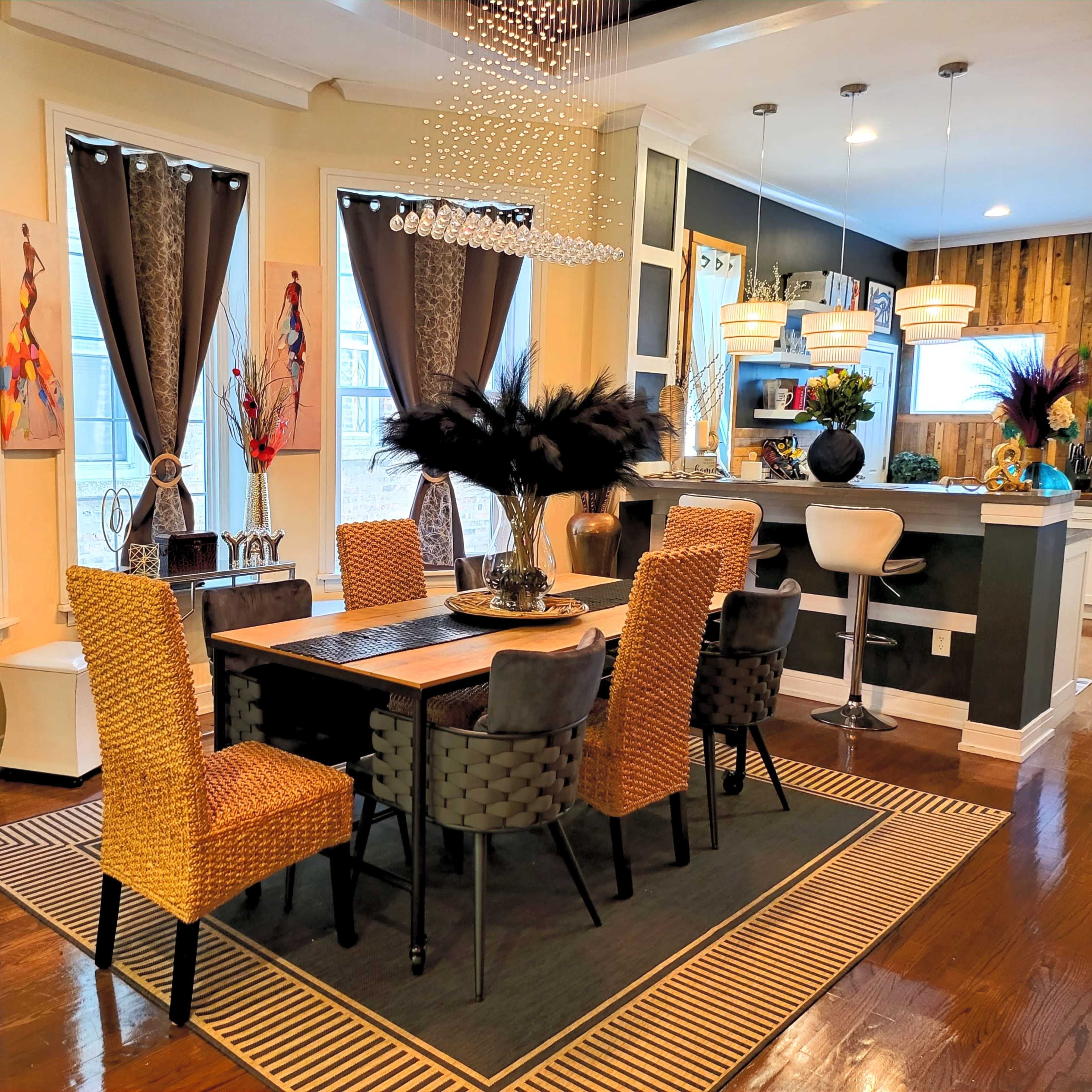 A dining area features a wooden table surrounded by woven and upholstered chairs, with large windows draped in dark curtains and a modern kitchen visible in the background.