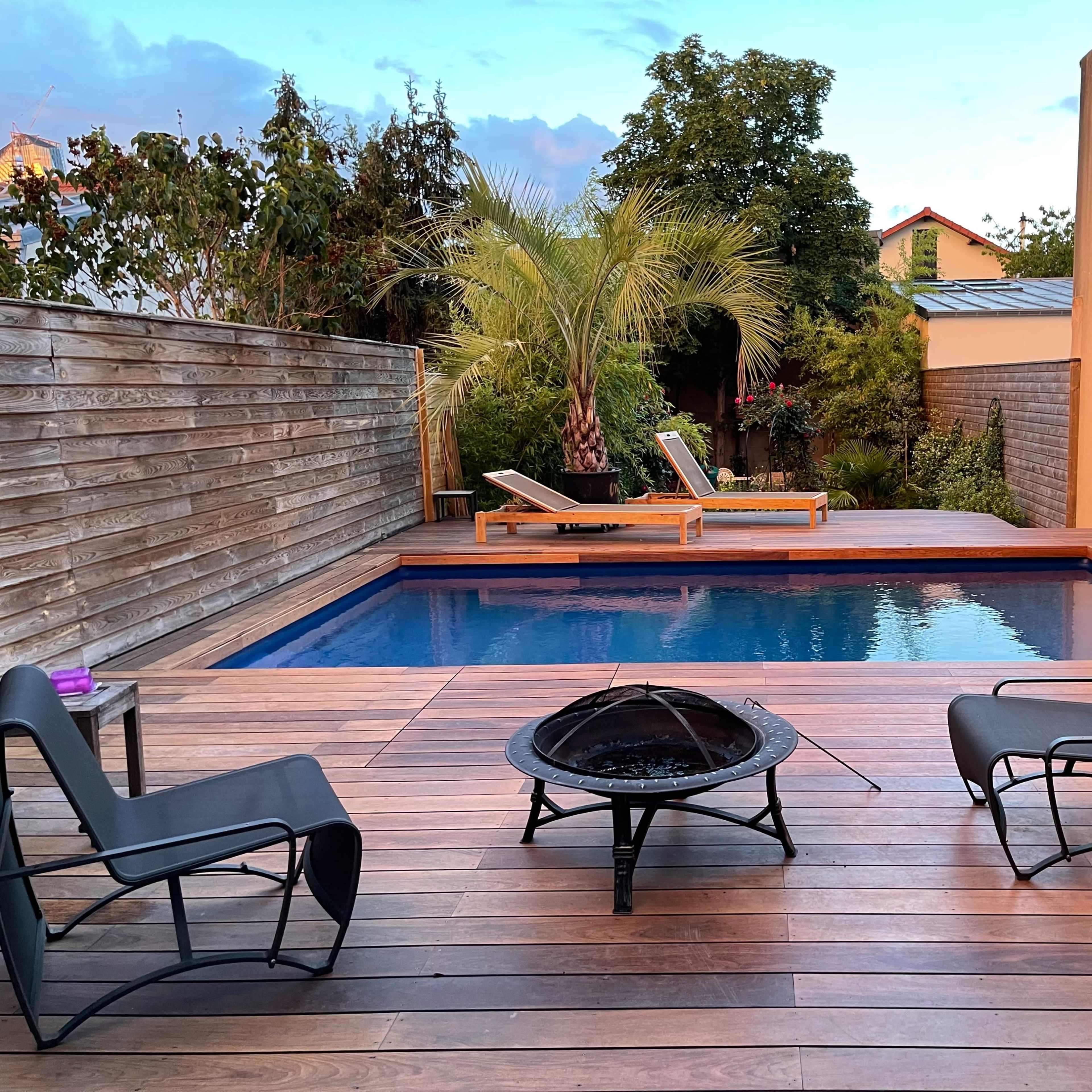 A backyard area featuring a small pool surrounded by wooden decking, two lounge chairs, and a fire pit.