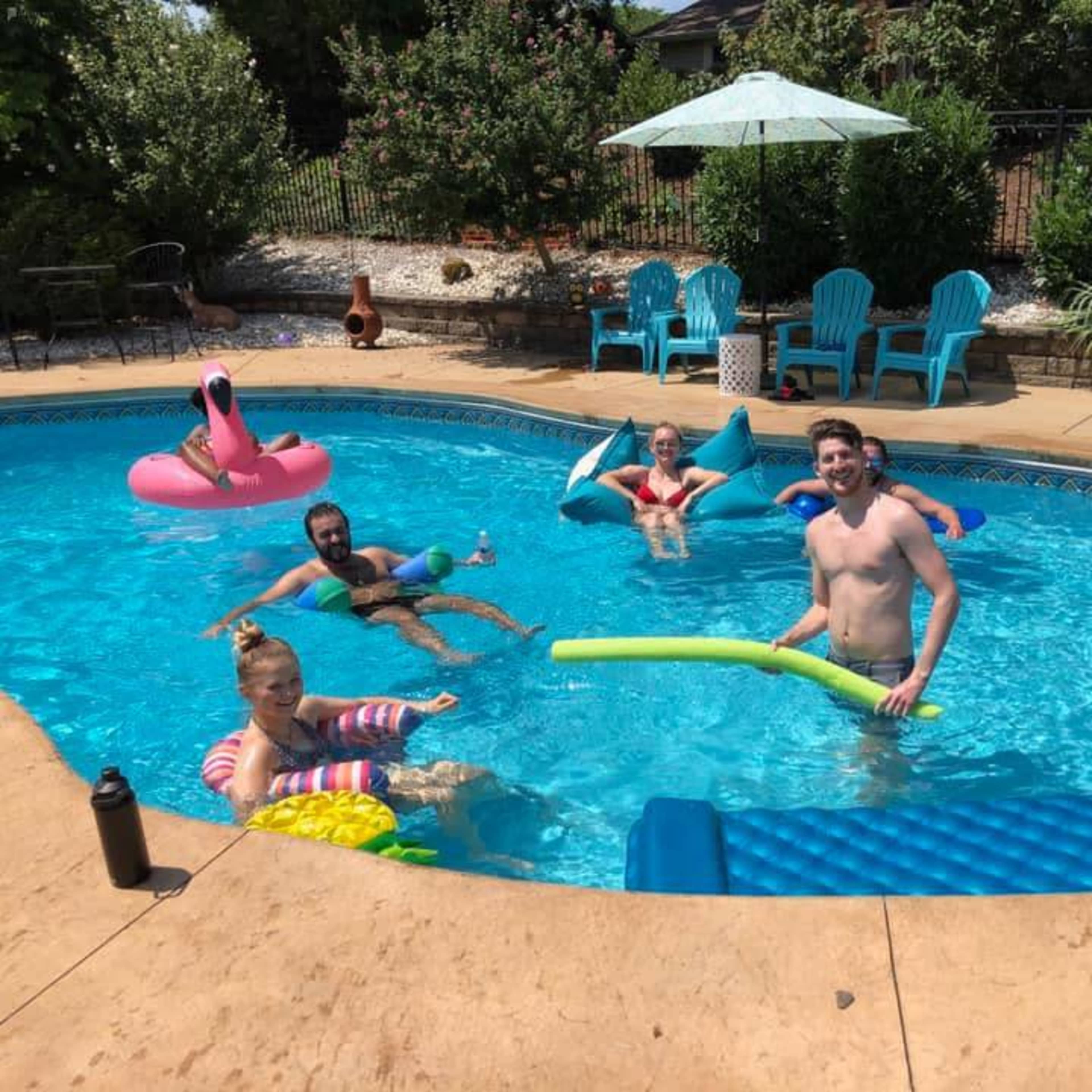 People are relaxing in a swimming pool surrounded by lounge chairs and a sun umbrella.
