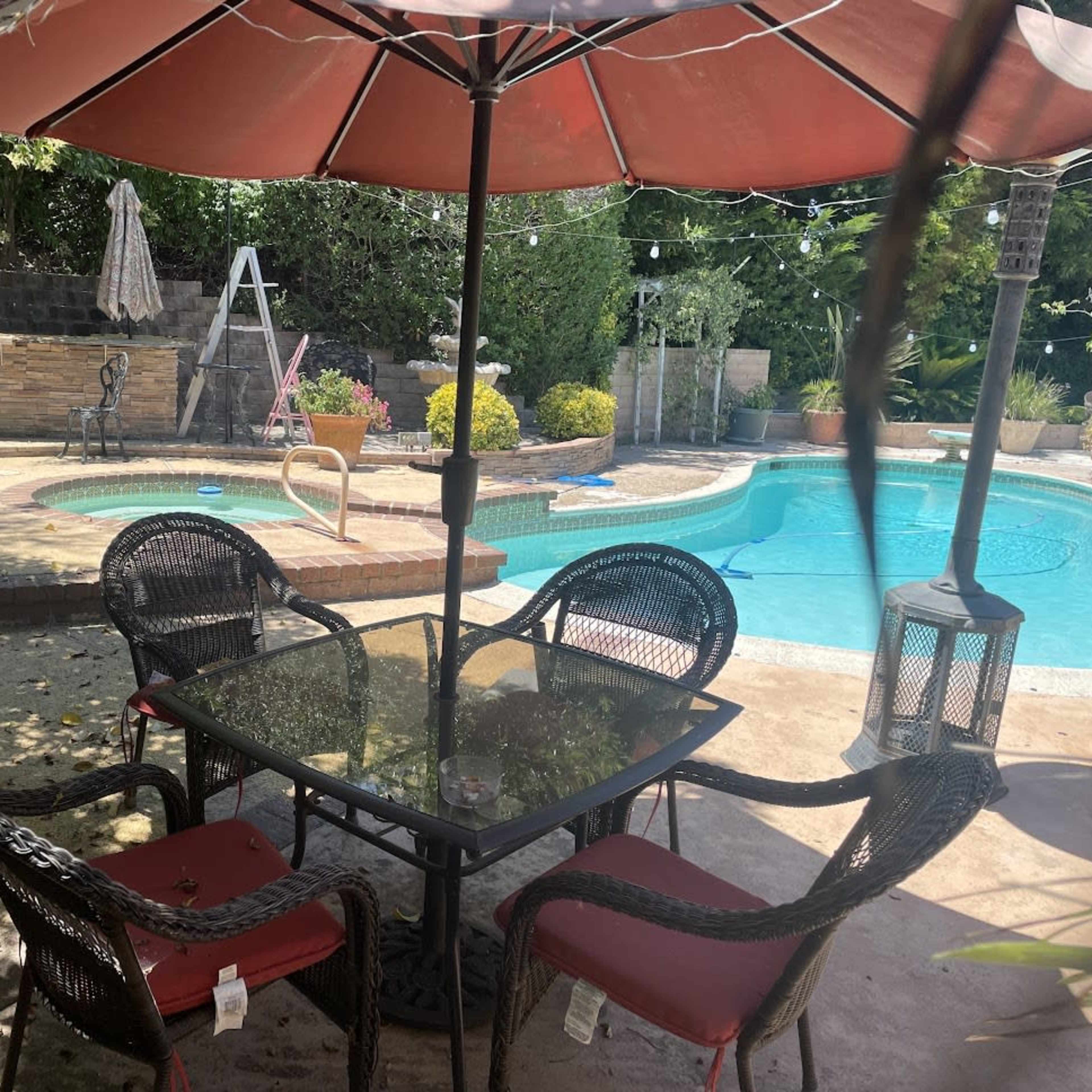 A patio area features a glass table and four chairs under a red umbrella, with a swimming pool and landscaped greenery in the background.