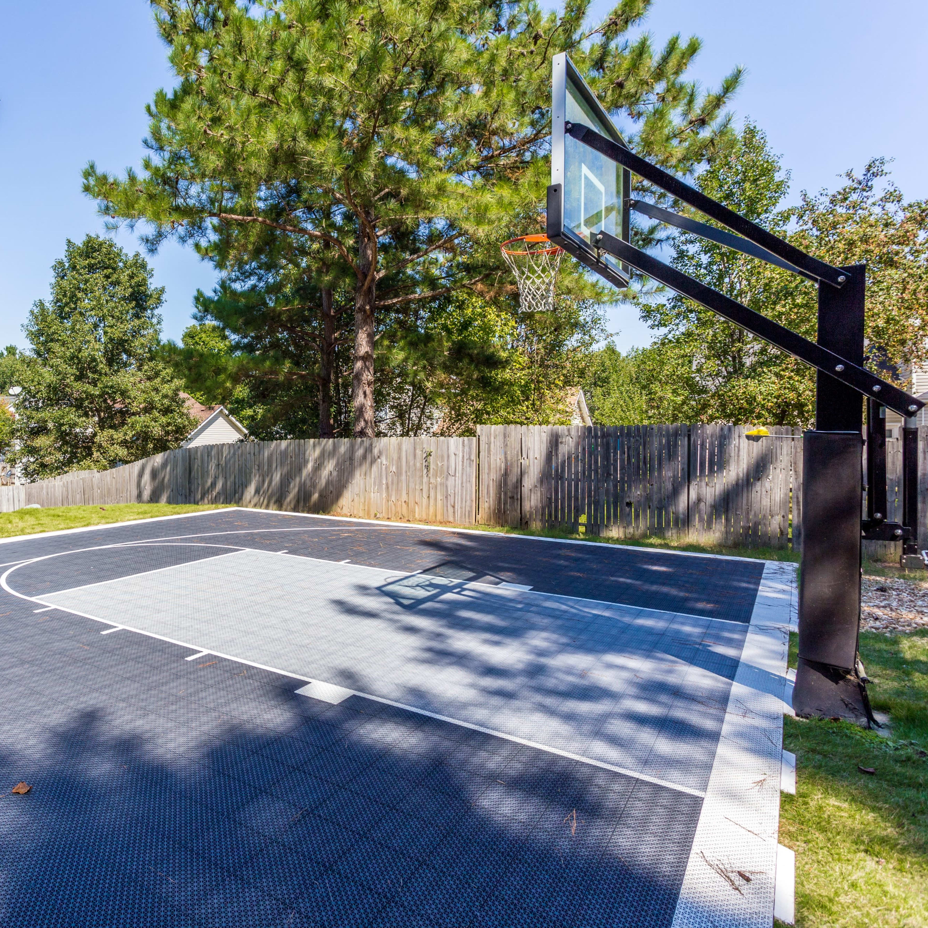 An outdoor basketball court surrounded by a wooden fence and trees, with a basketball hoop mounted on a black support structure.