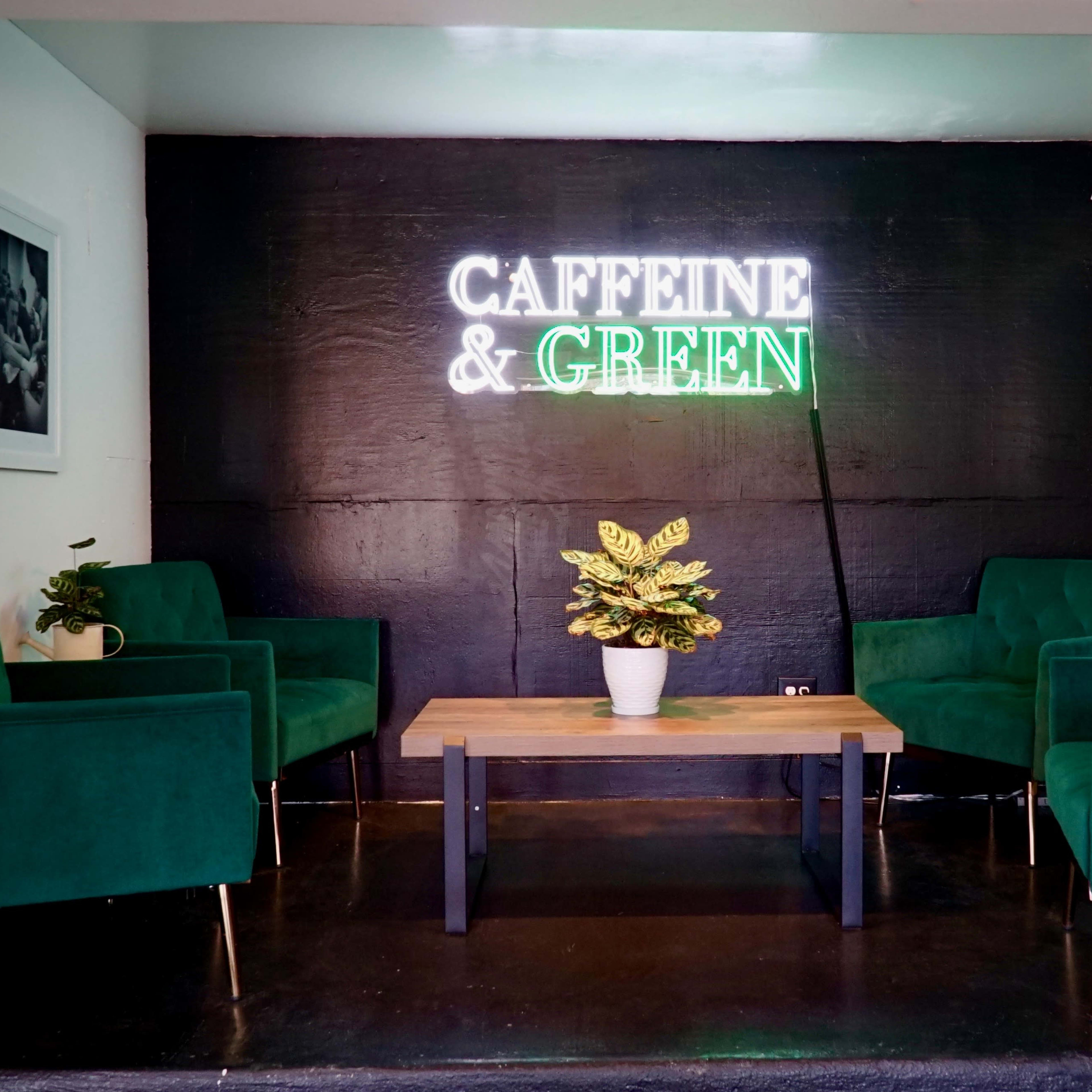 A seating area with four green chairs and a wooden table, illuminated by a neon sign that reads "CAFFEINE & GREEN."