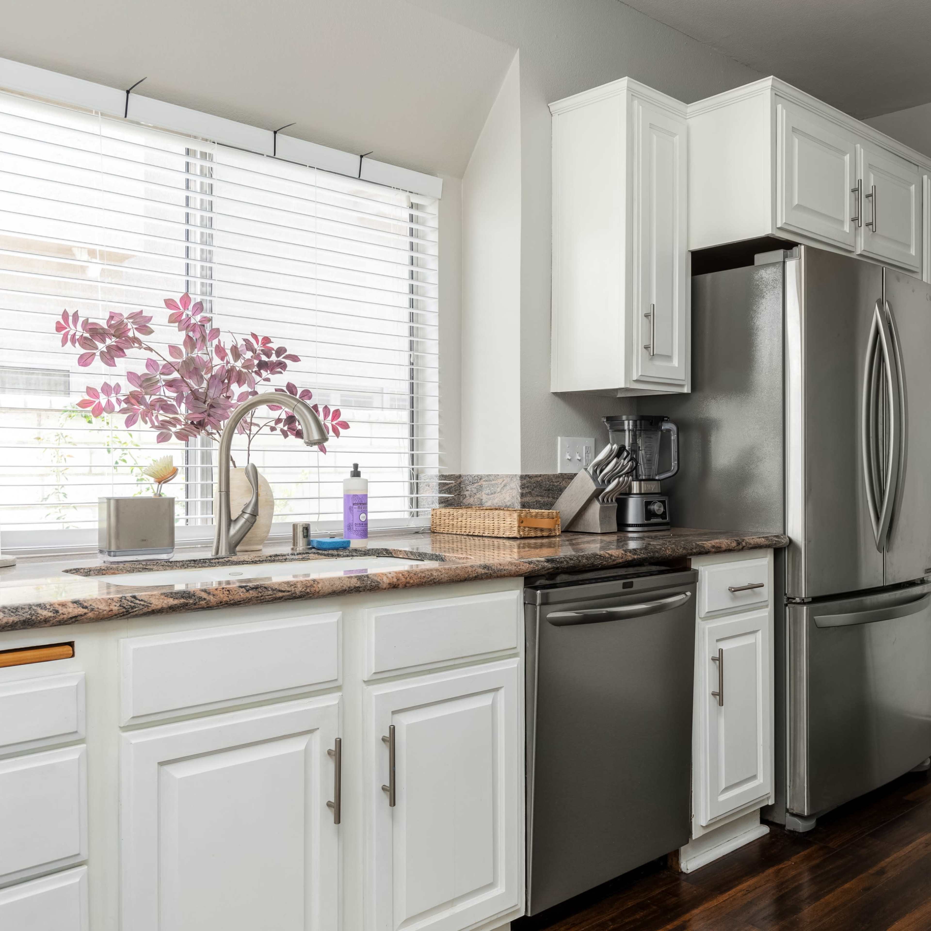 A modern kitchen features white cabinetry, a stainless steel refrigerator, and a granite countertop with a window adorned by a vase of purple flowers.