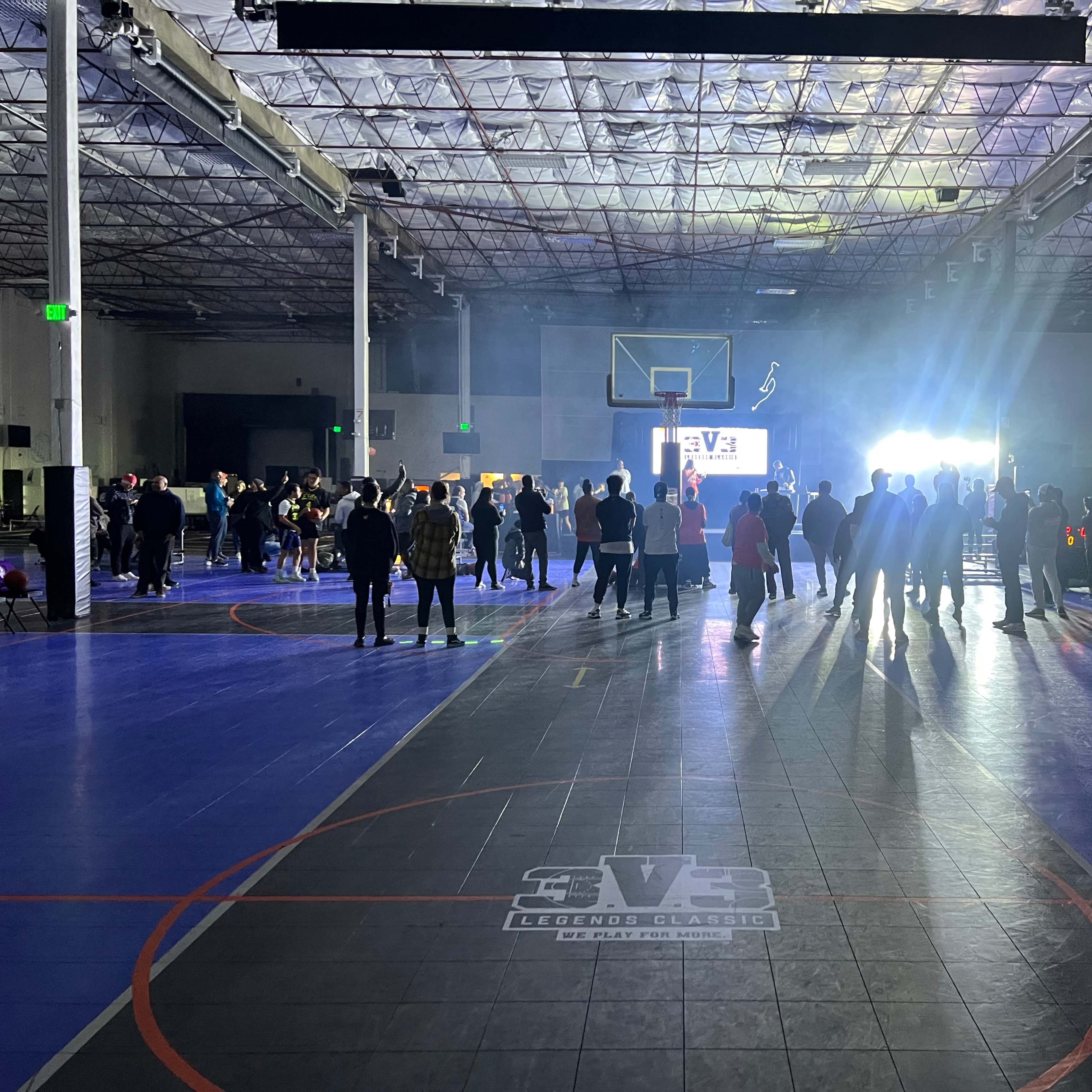 A crowd gathers in a dimly lit basketball gymnasium with a illuminated stage at one end.