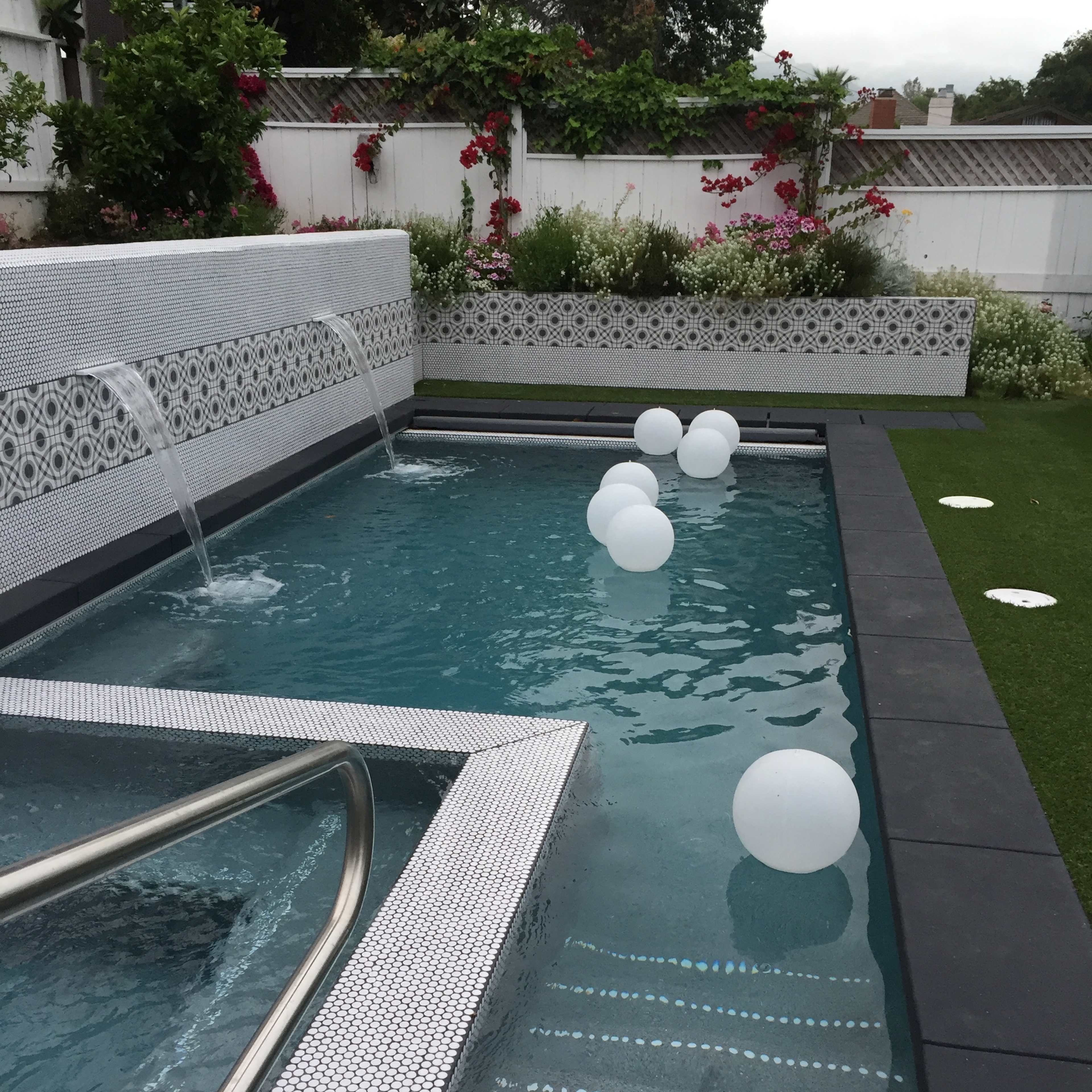 A modern pool with steps leading down into the water, flanked by decorative tiles and three white floating spheres, surrounded by greenery and flowers.
