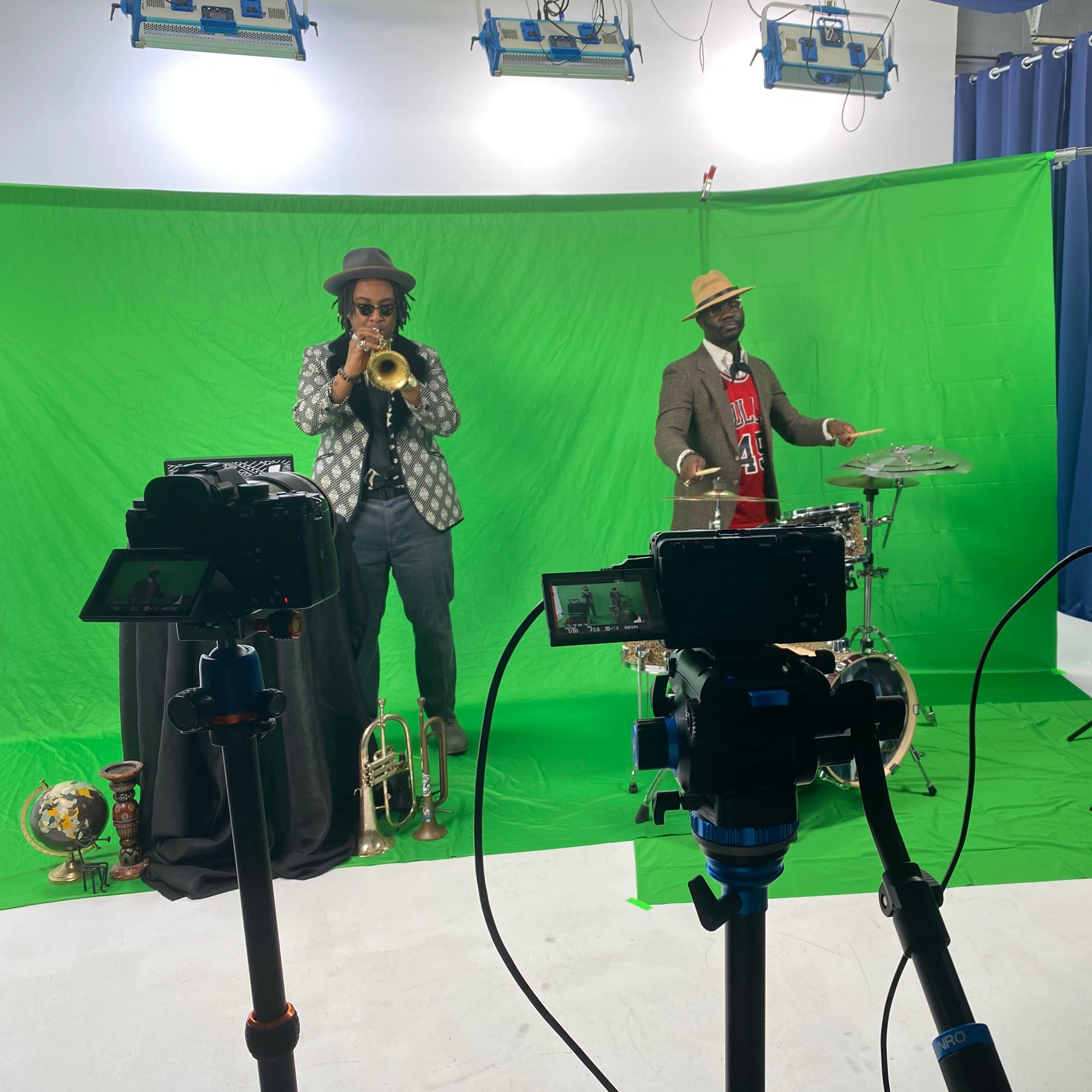 Two musicians perform in front of a green screen, one playing the trumpet and the other playing the drums, while several cameras are positioned in the foreground.