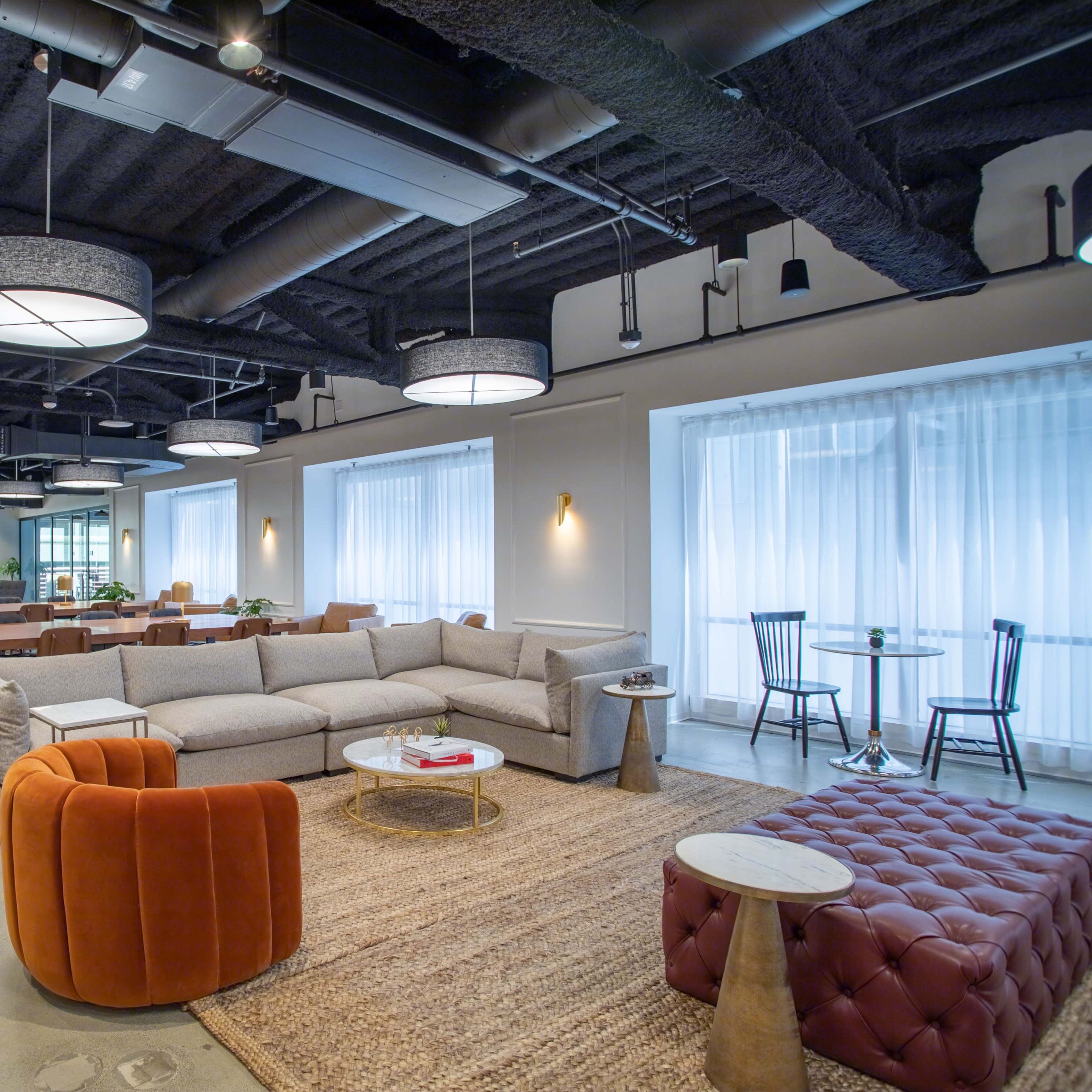A modern lounge area featuring a gray sectional sofa, orange accent chairs, and a variety of tables under circular ceiling lights.