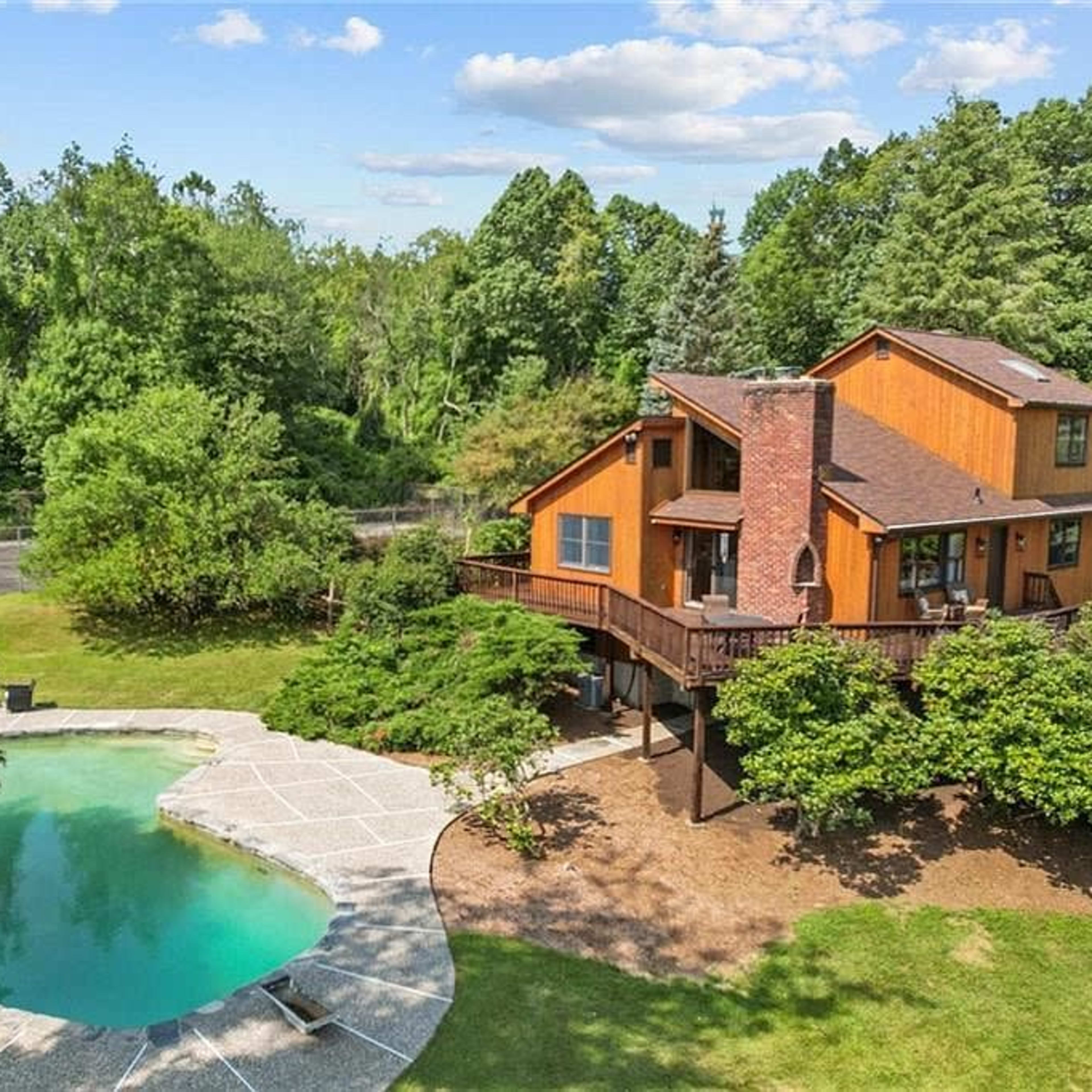 A two-story wooden house with large windows overlooks a curved swimming pool surrounded by landscaped greenery.