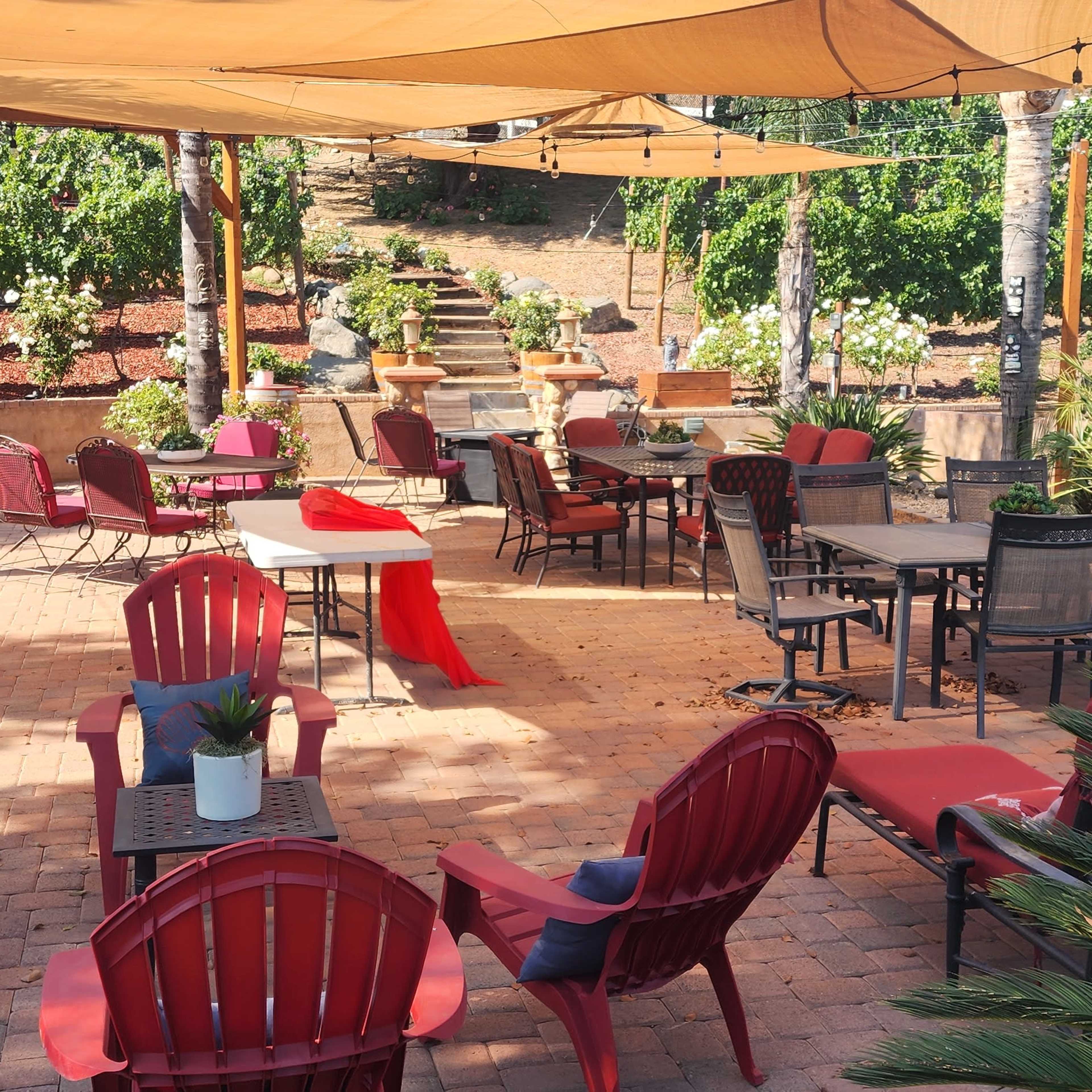 An outdoor seating area with red and black furniture, shaded by fabric canopies, surrounded by greenery and decorative planters.