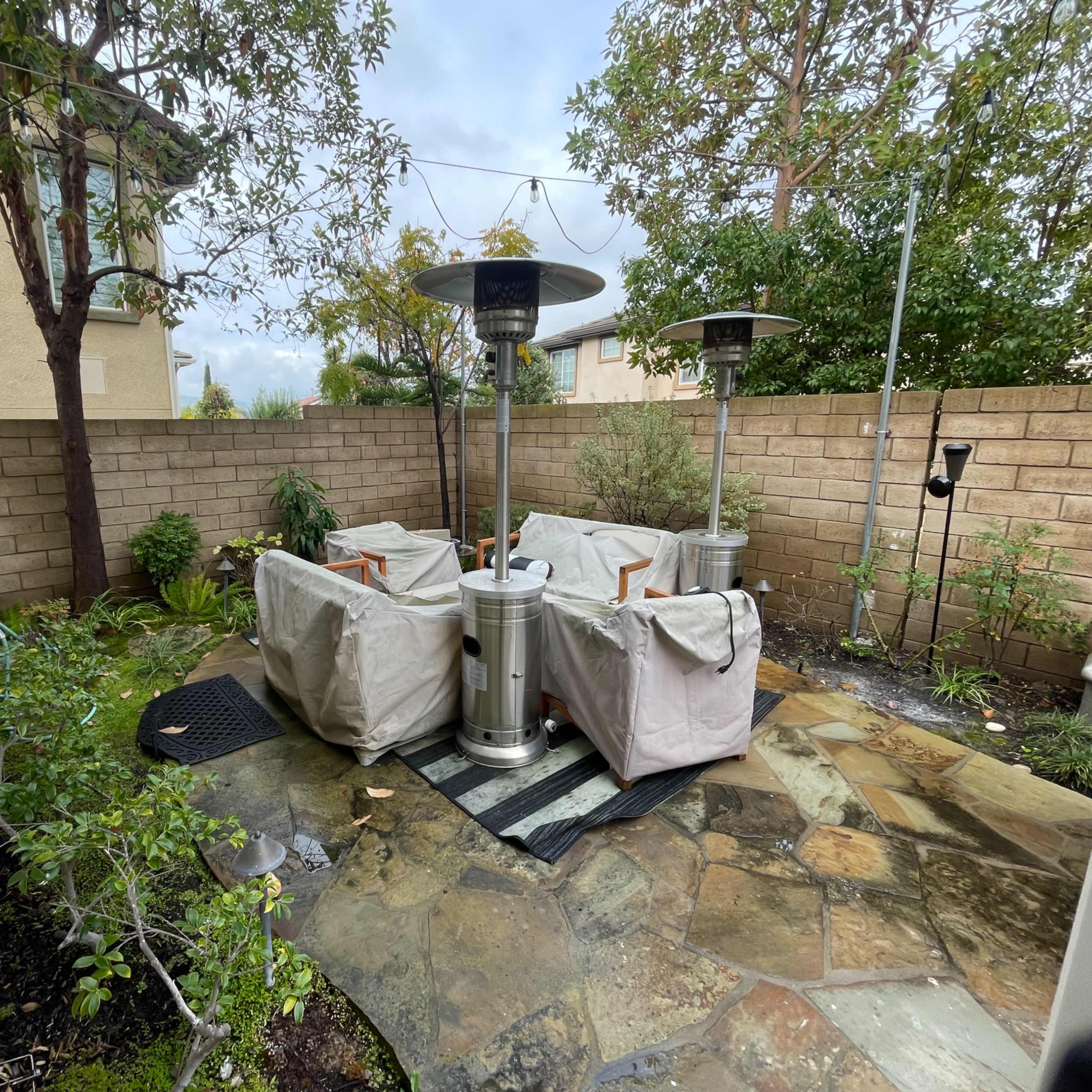 A patio area enclosed by a stone wall, featuring four covered patio heaters arranged around a stone pathway amidst greenery.