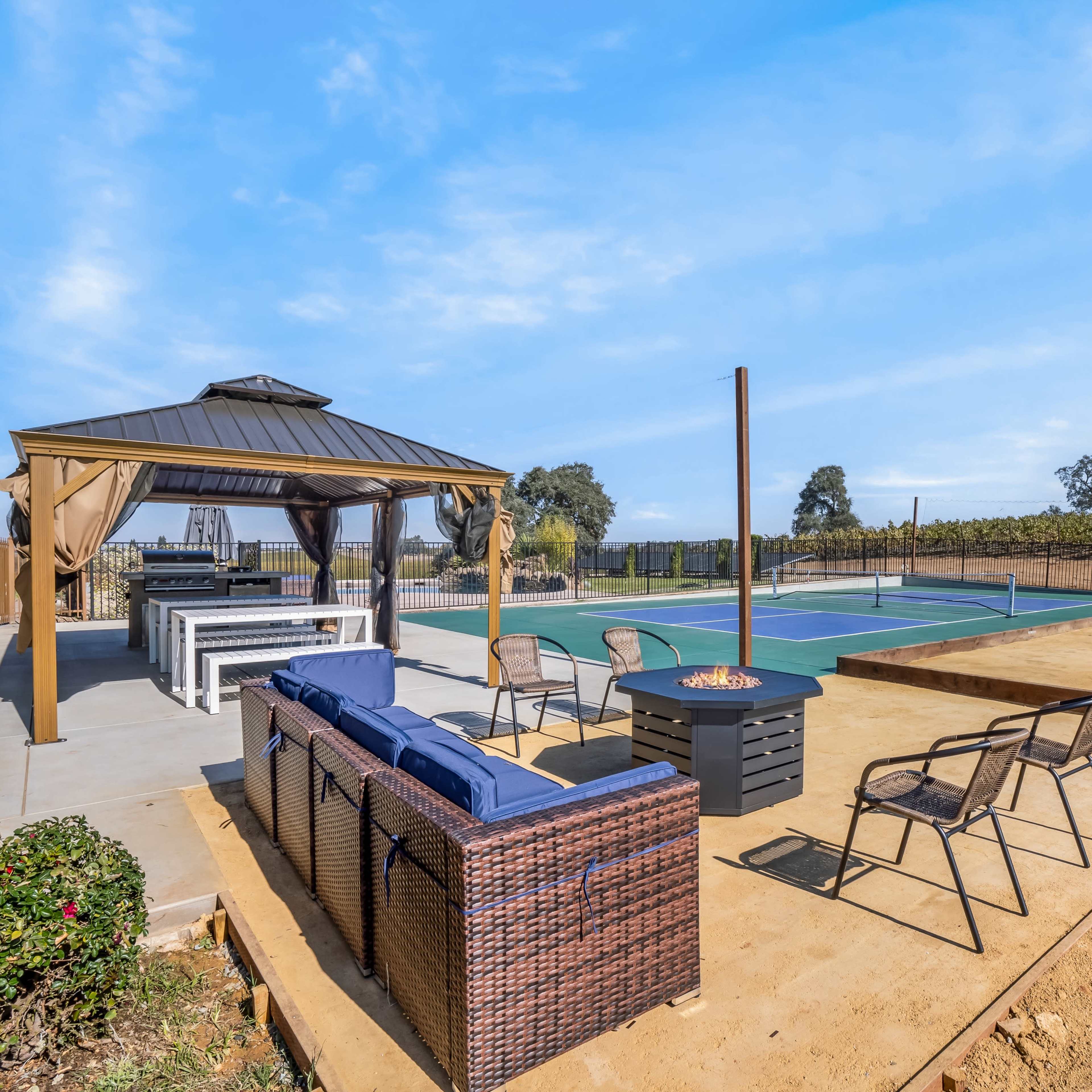 A patio area with seating and a fire pit beside a tennis court, surrounded by vineyards.
