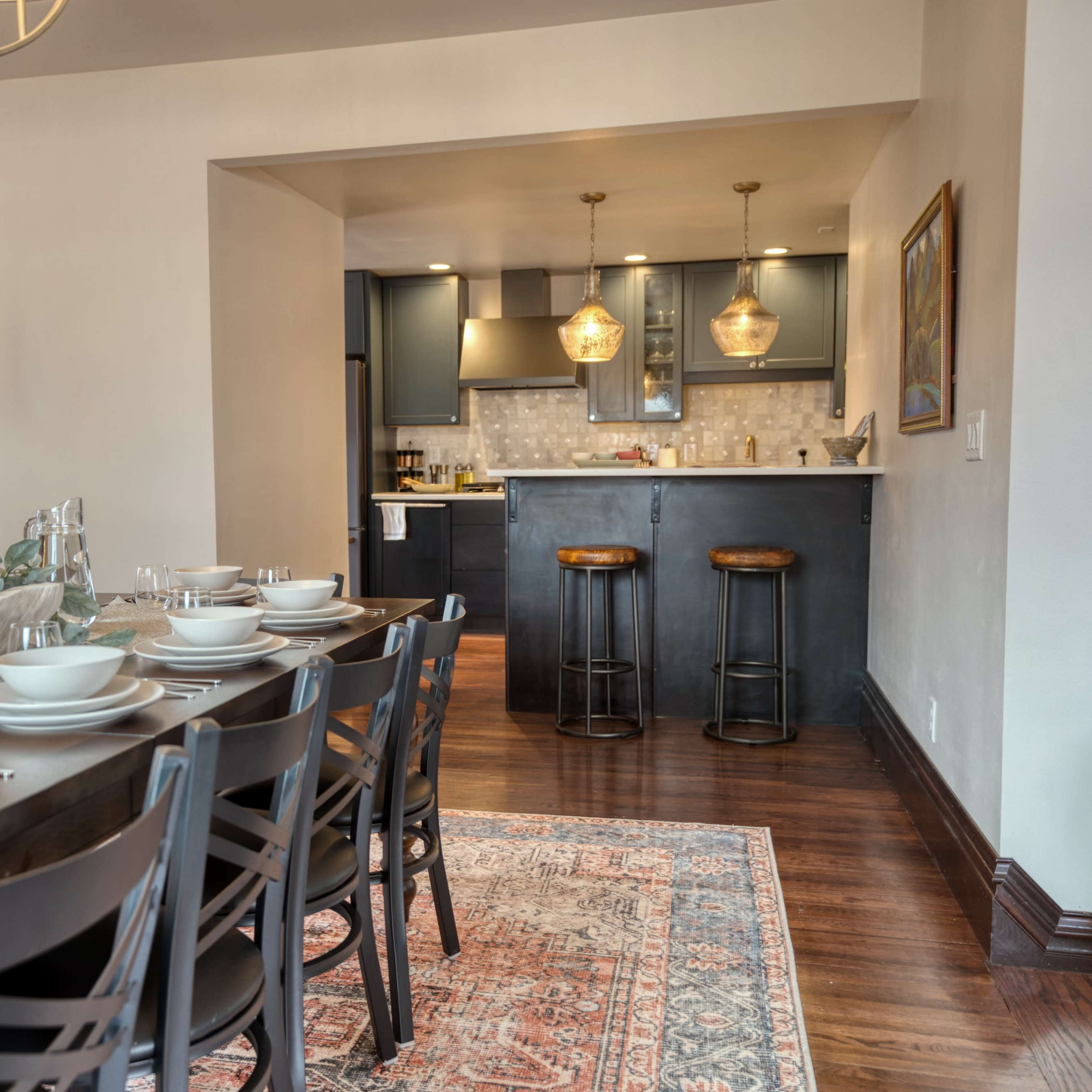 A dining area features a long table set with dishes, leading to a kitchen with dark cabinets and bar stools.
