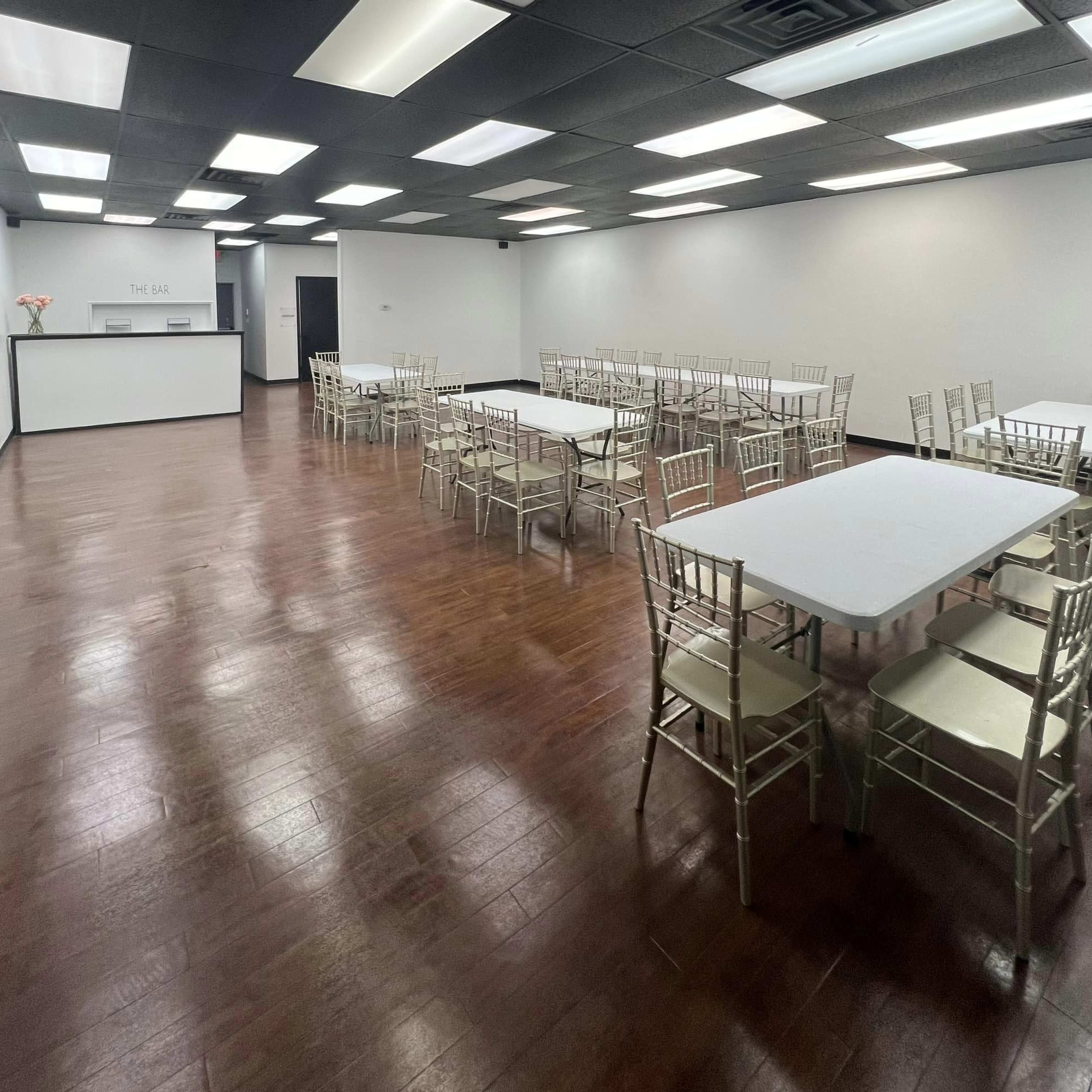 A spacious event room with multiple rectangular tables and chiavari chairs arranged in two sections, with a reception area visible in the background.