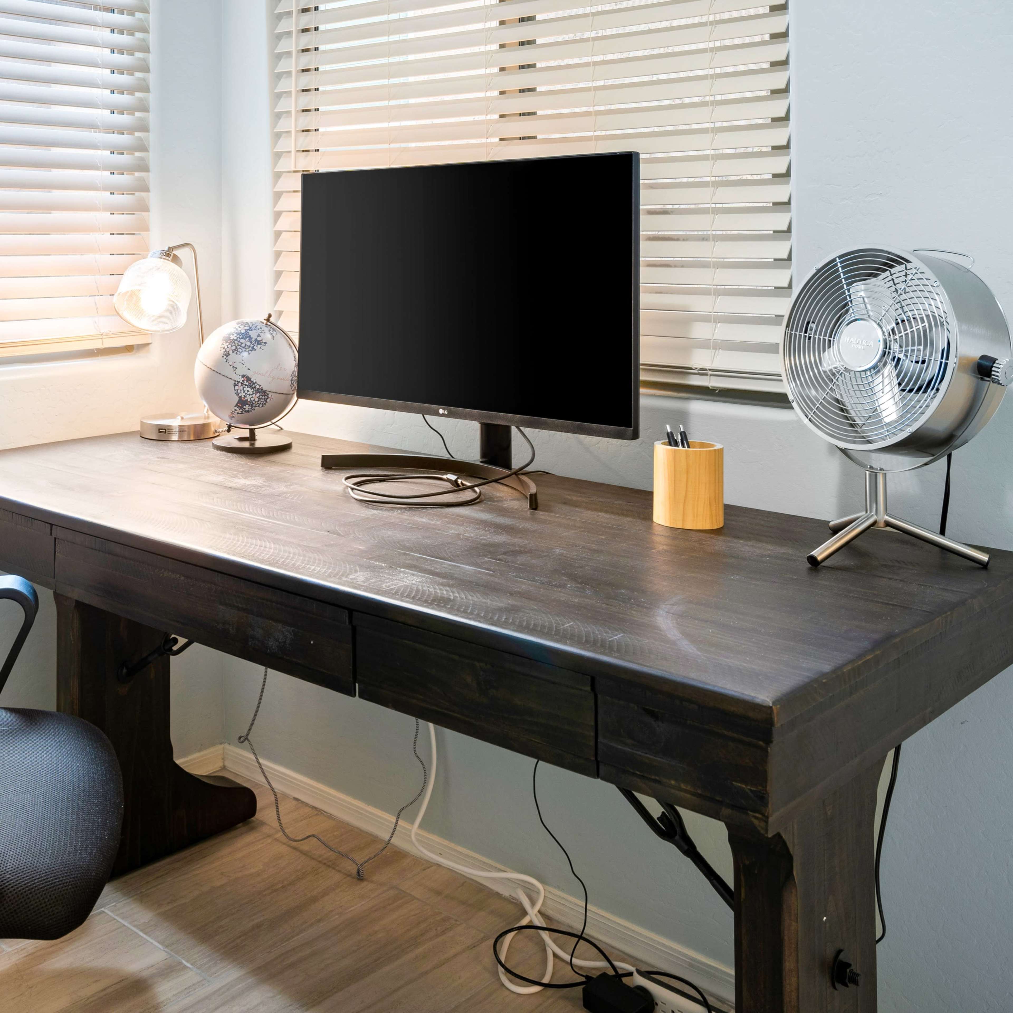 A wooden desk is positioned next to a window with blinds, featuring a monitor, a globe, a fan, and a black office chair.