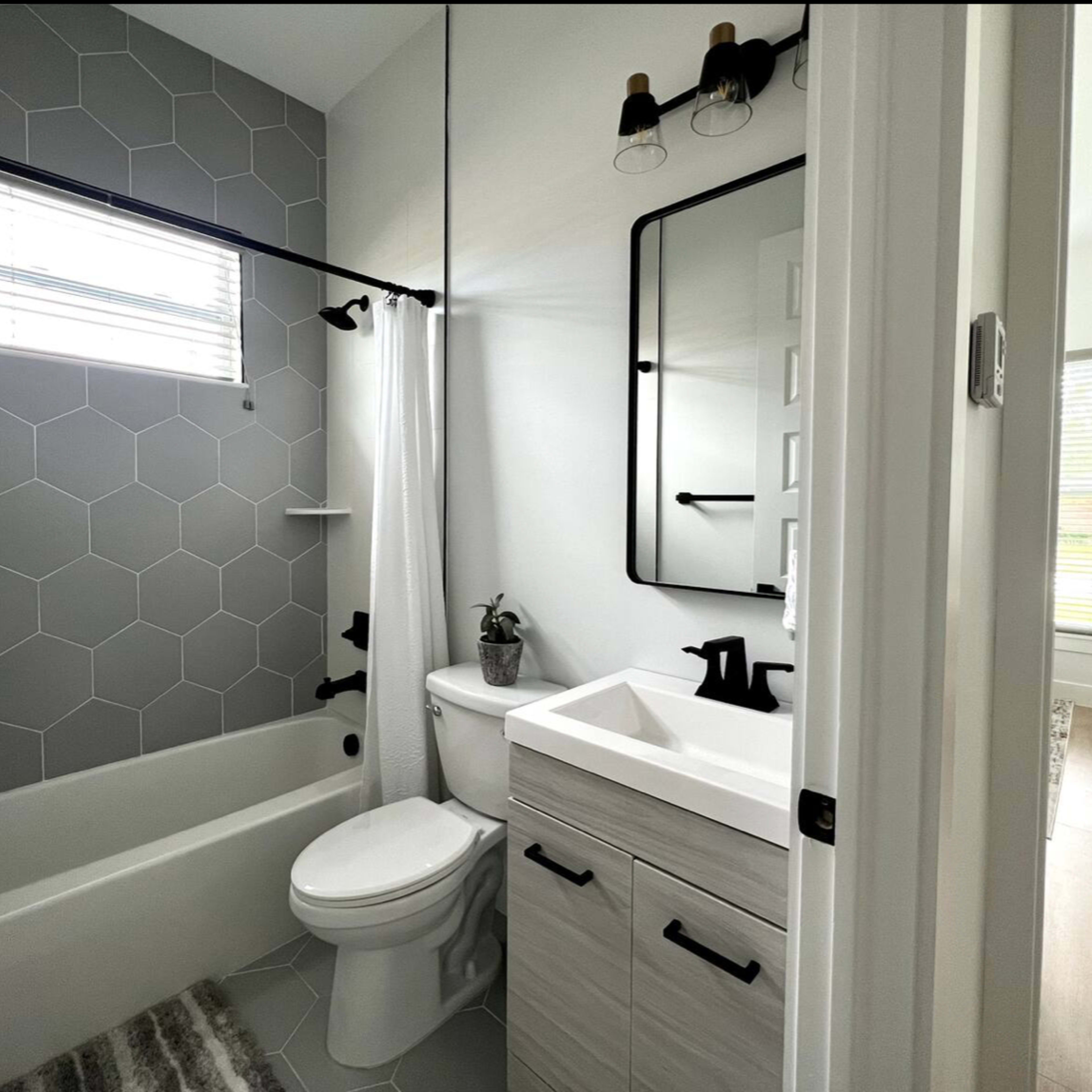 A modern bathroom featuring a hexagon-tiled wall, a bathtub with a shower, a toilet, and a sink with a rectangular mirror.