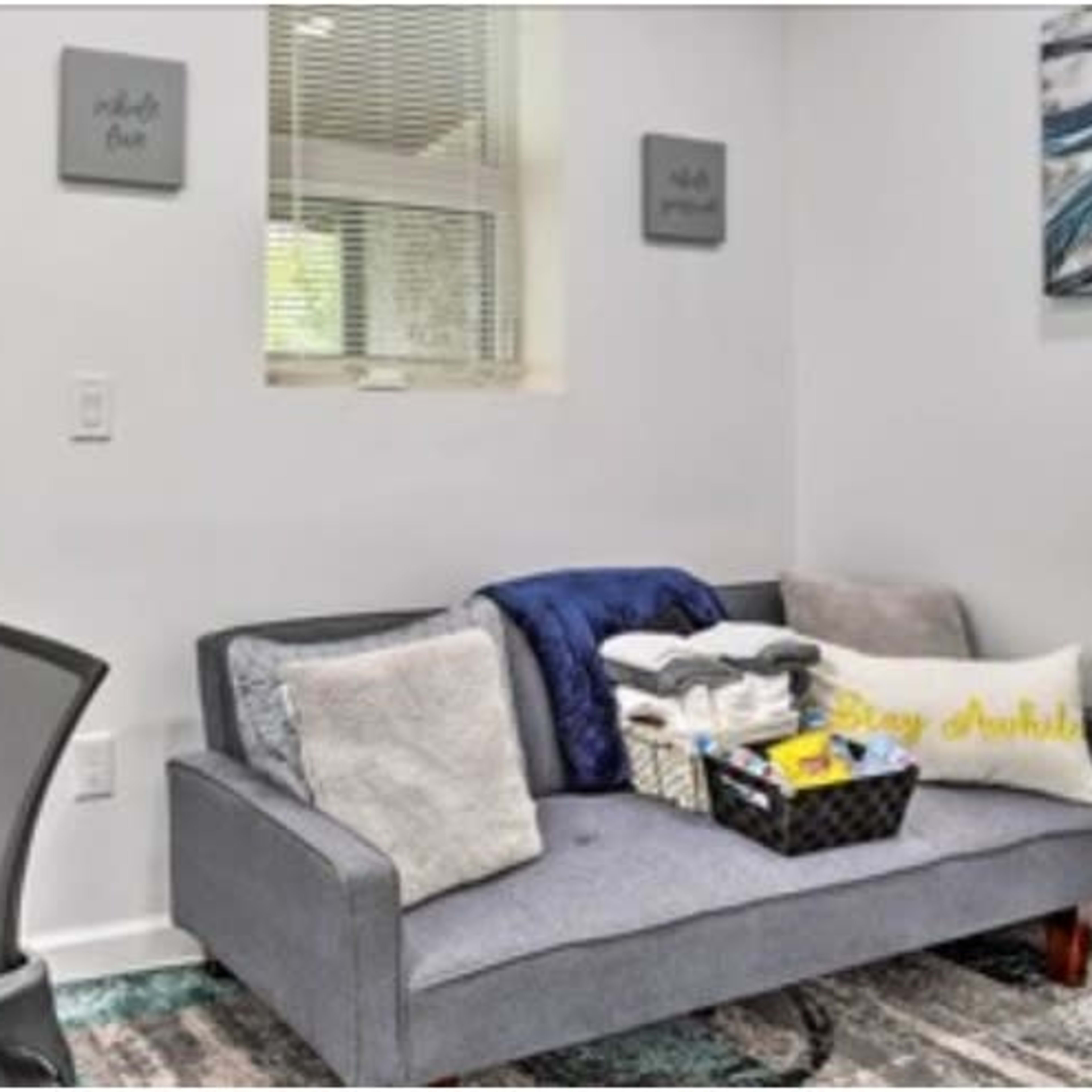 A small office space with a desk, chair, and a gray couch adorned with blankets and a basket of items against a white wall.