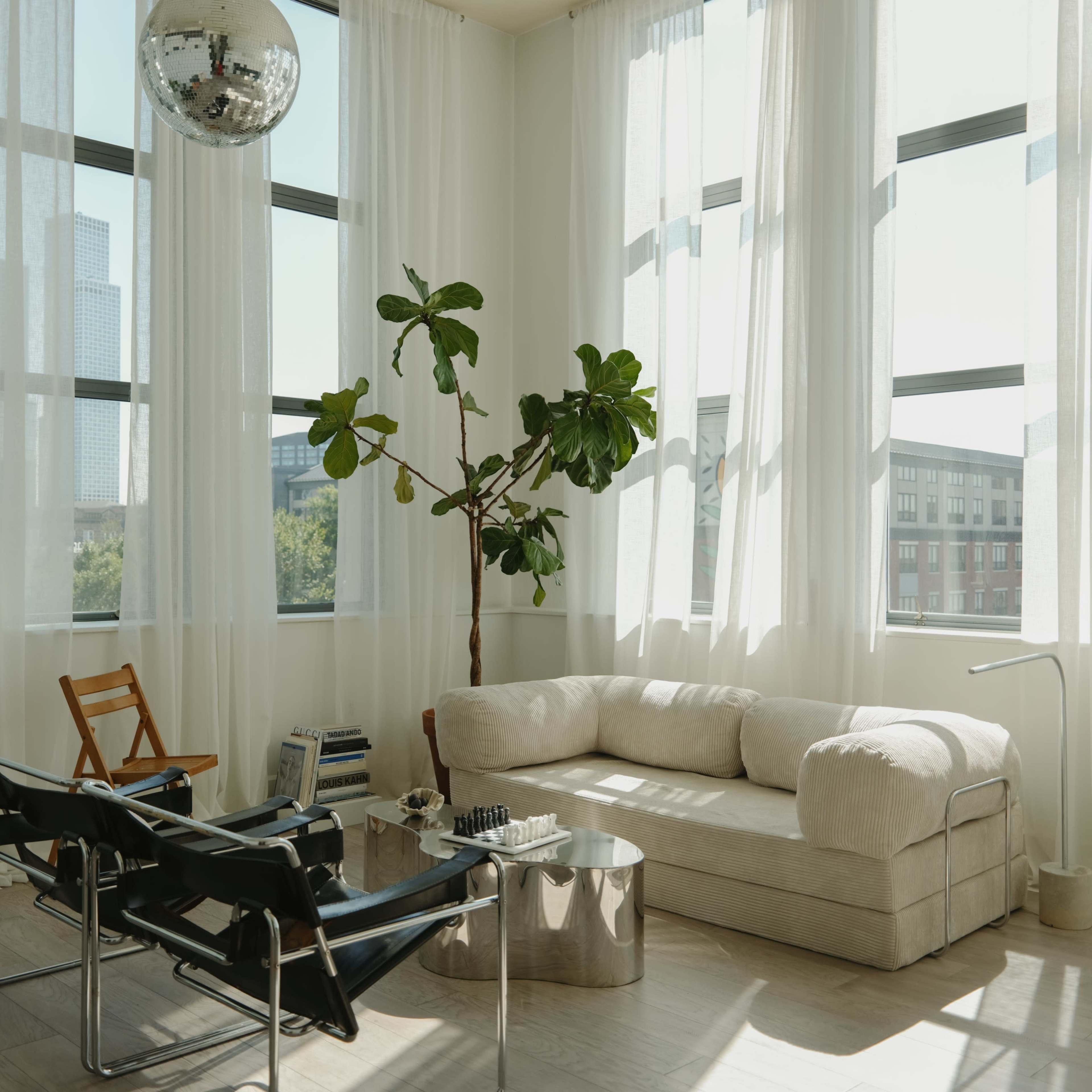 A spacious living room features large windows with sheer curtains, a comfortable sofa, two black chairs, a coffee table, a potted plant, a guitar, and a disco ball hanging from the ceiling.