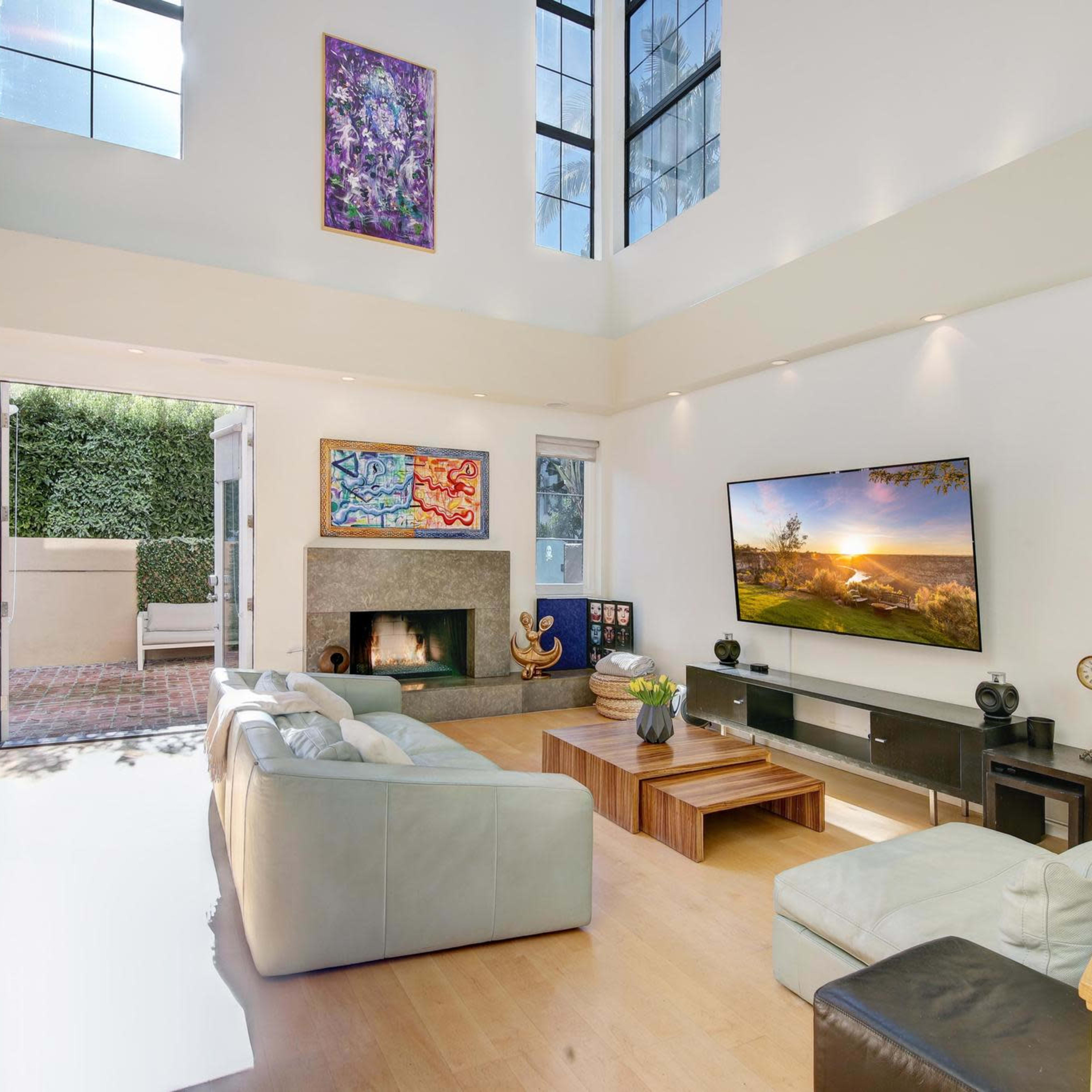 A modern living room with a large TV mounted on the wall, a fireplace, and large windows allowing natural light to enter.