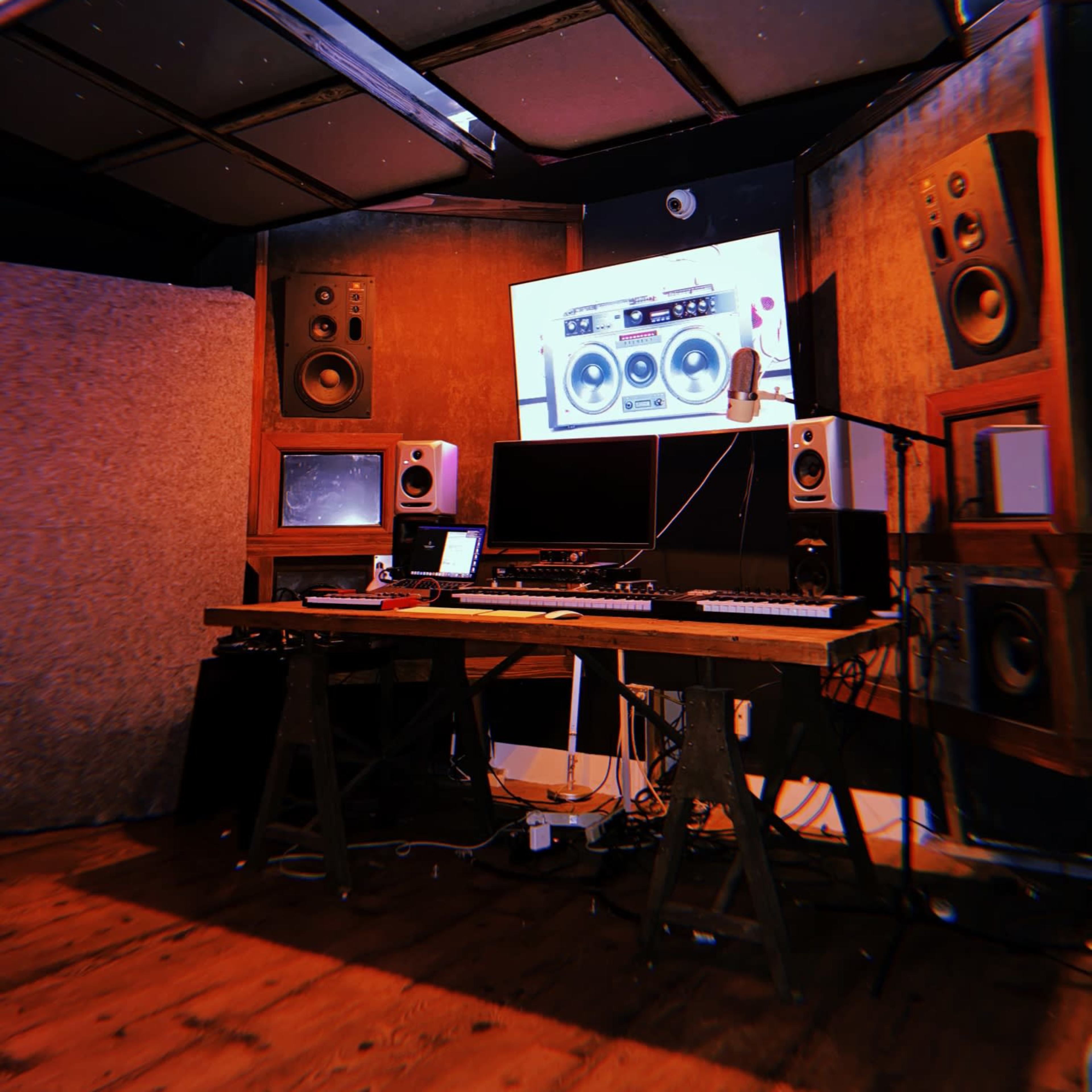 A music studio is shown with wooden flooring, large speakers, a computer, and a keyboard setup in front of a wall-mounted screen.