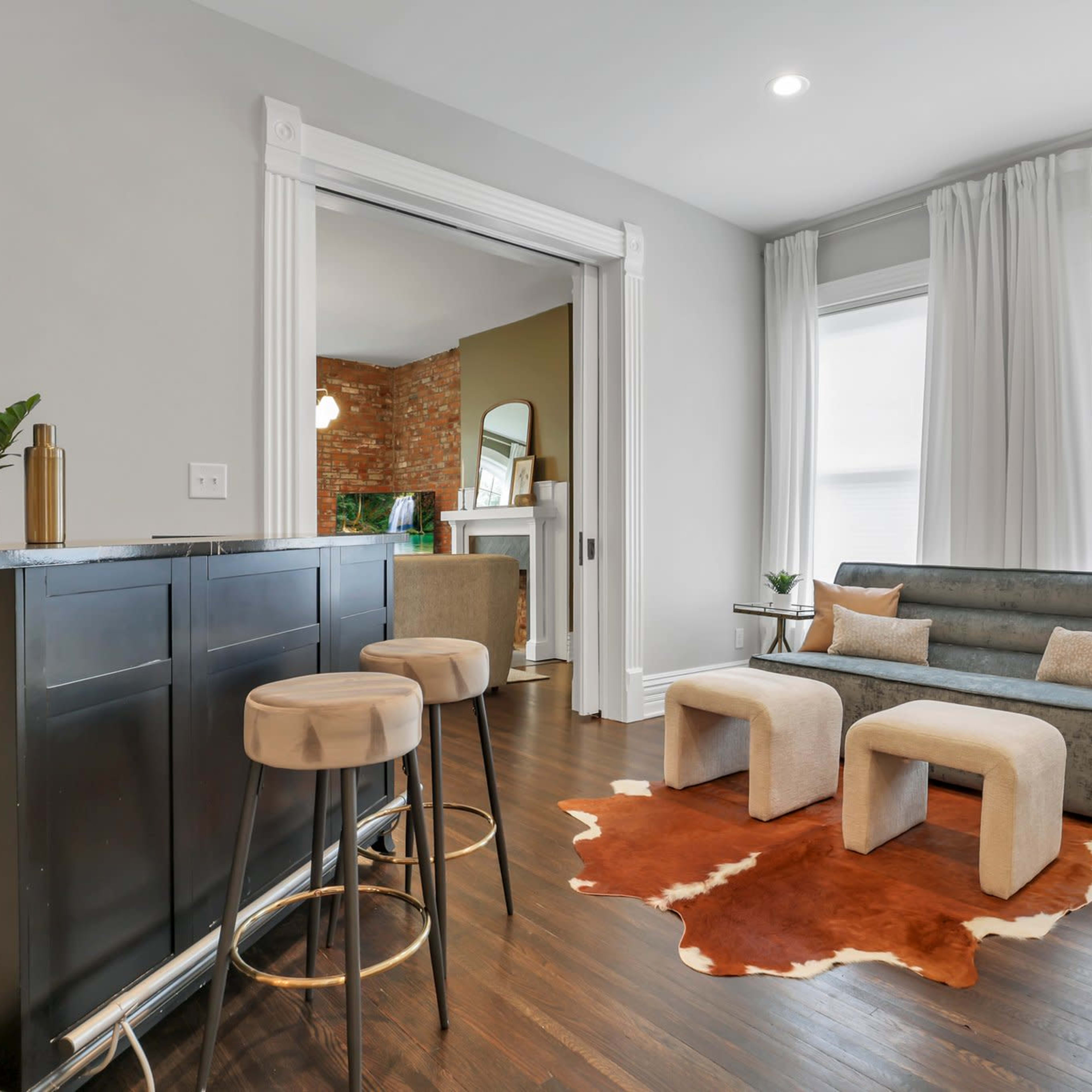 A modern living space with a small bar area, a gray sofa, two light-colored stools, and a cowhide rug on dark hardwood flooring.