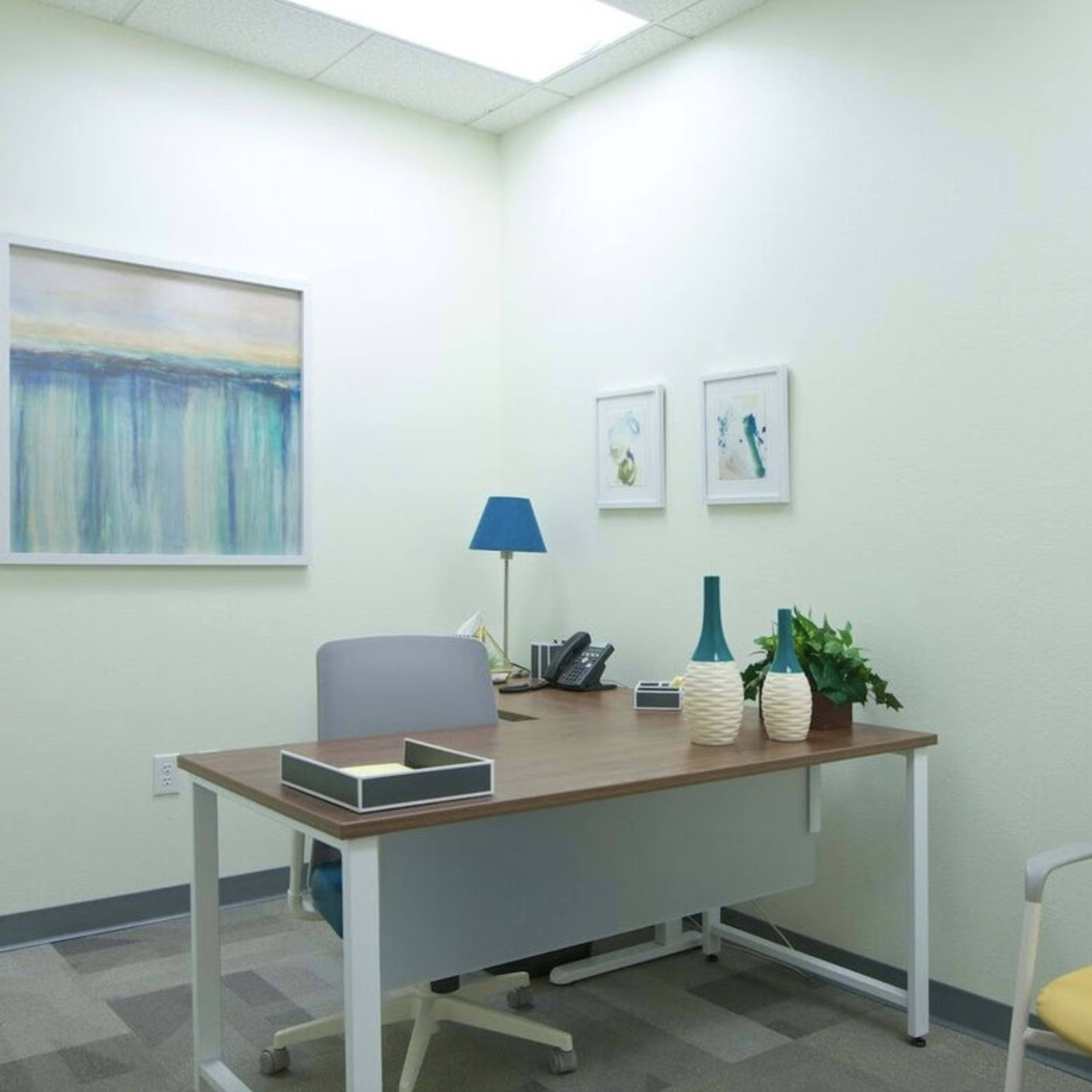 A small office space with a wooden desk, two chairs, a telephone, and decorative items including a lamp and artwork on the walls.