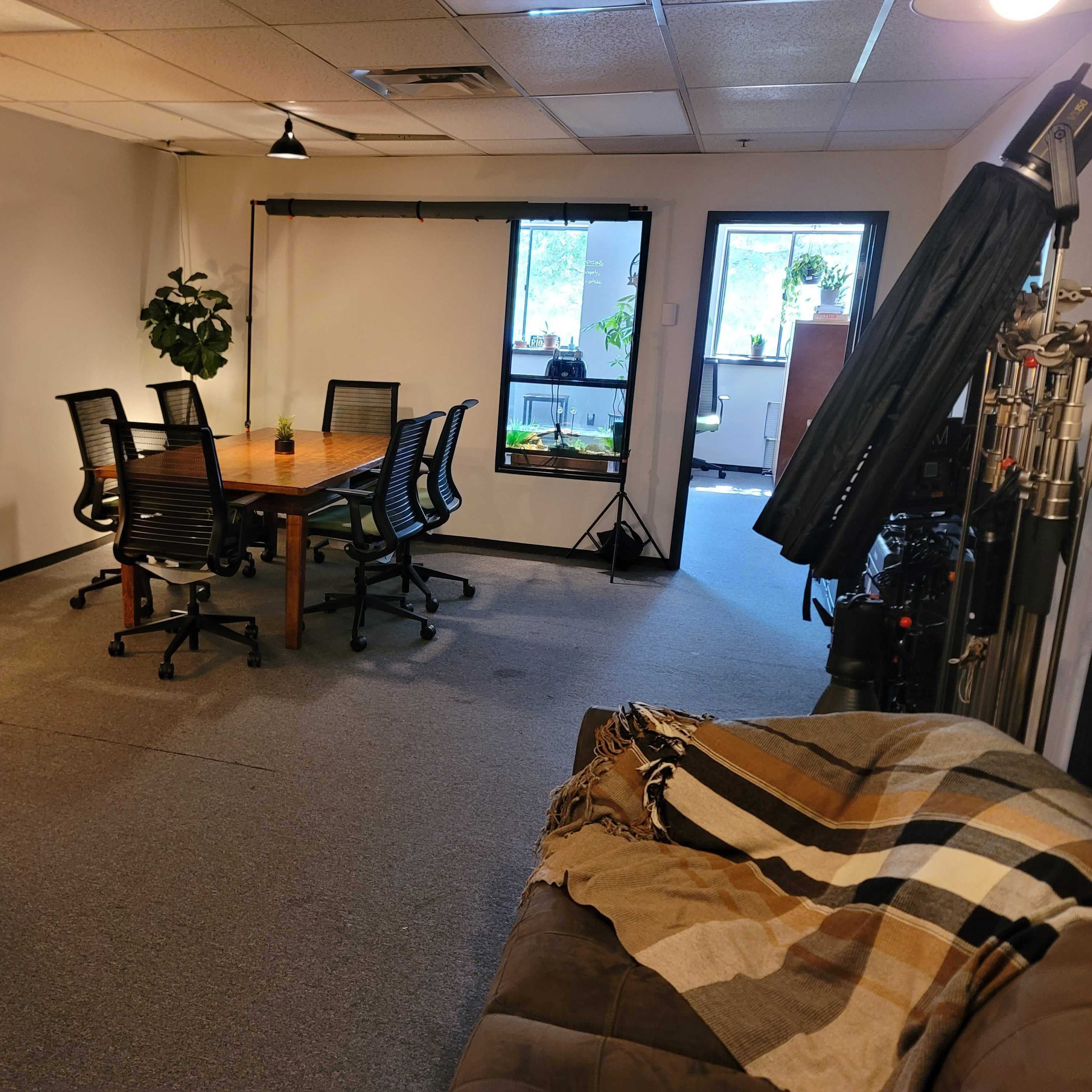 An office space featuring a wooden conference table surrounded by black chairs, a couch in the foreground, and large windows with plants nearby.