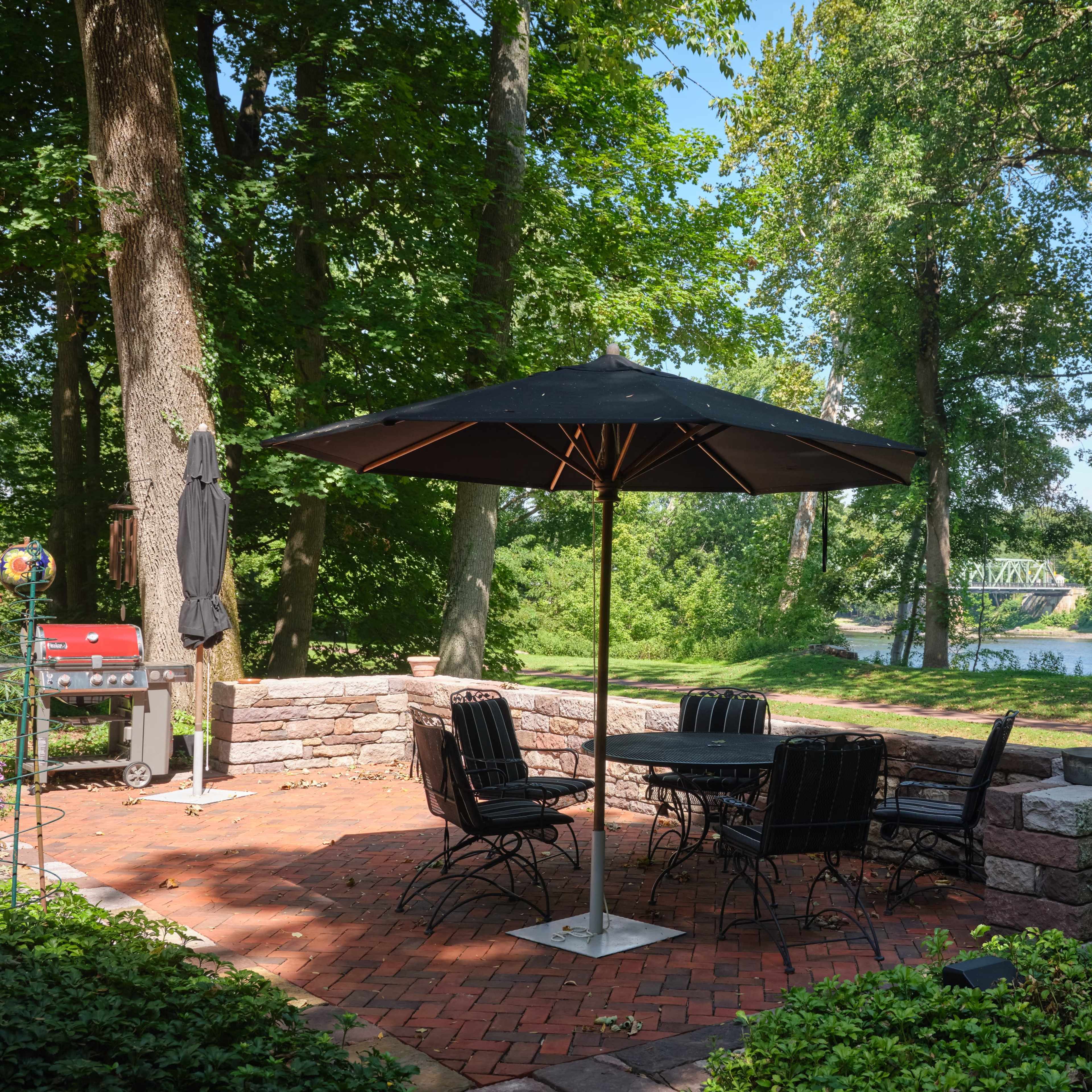 A brick patio with a table and chairs under a black umbrella, surrounded by trees and overlooking a river.