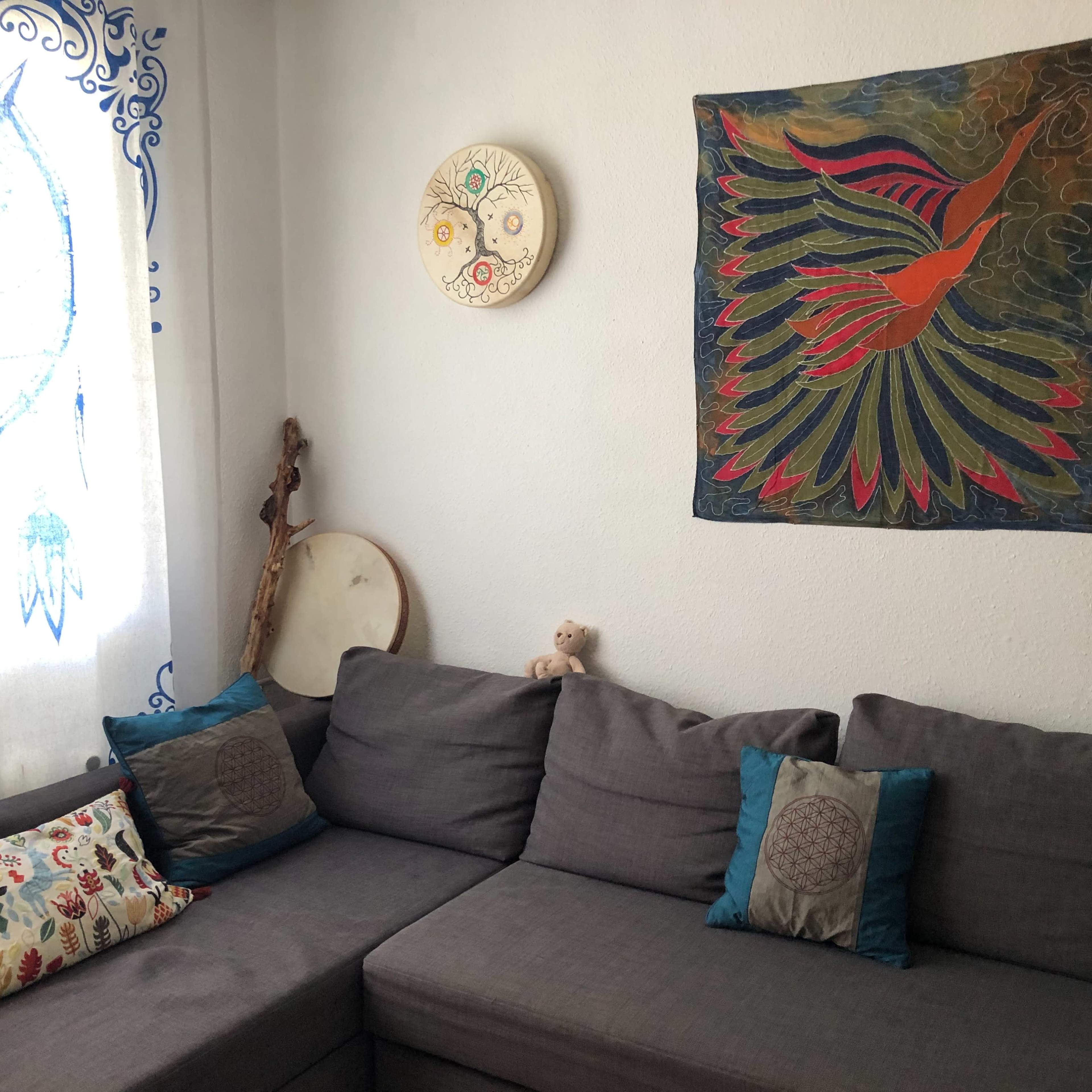 A cozy living room with a gray sectional sofa adorned with decorative pillows, a colorful wall hanging of a bird, and a window featuring a blue owl design.