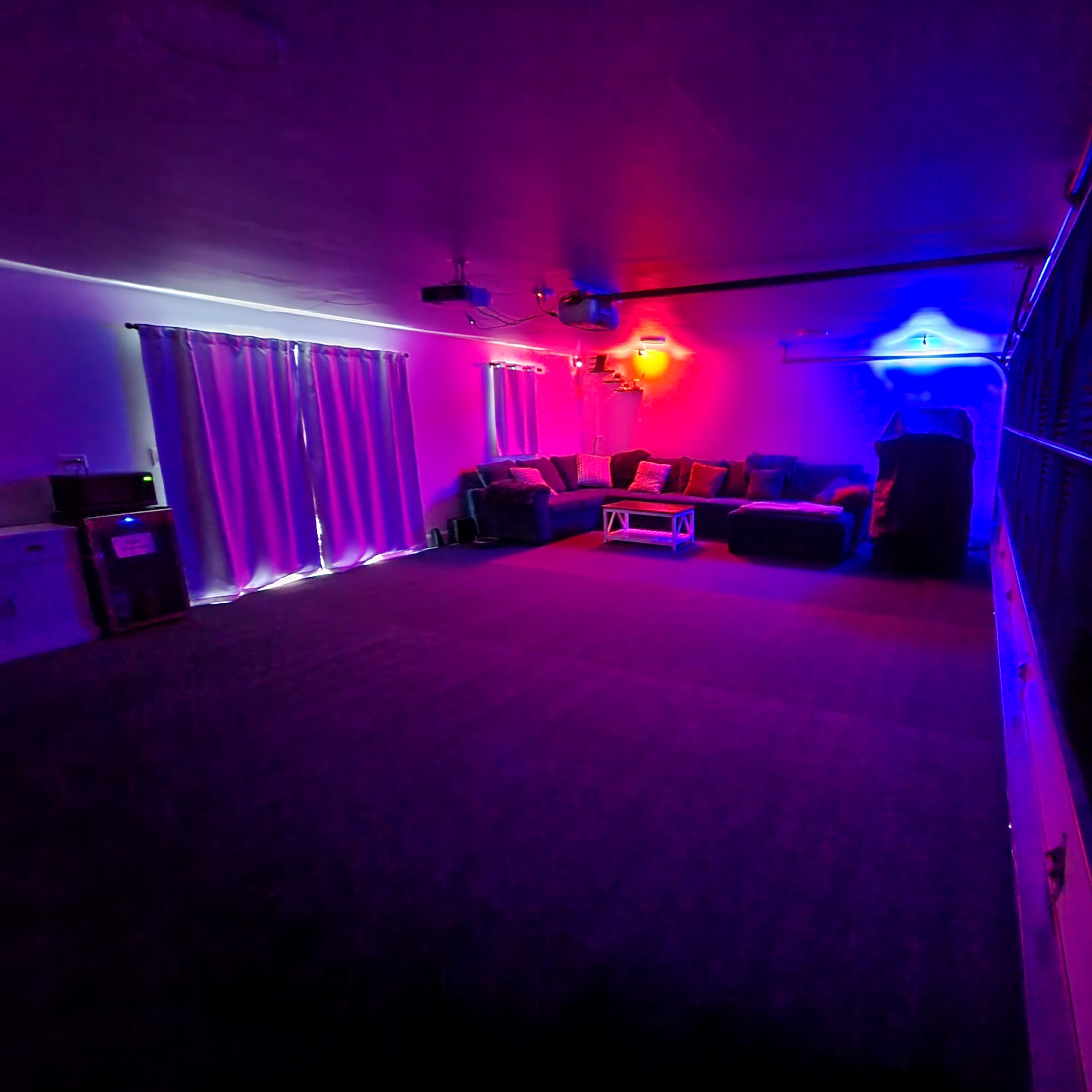 A spacious room features a sectional sofa with a coffee table, illuminated by colorful ambient lighting and a projector setup.