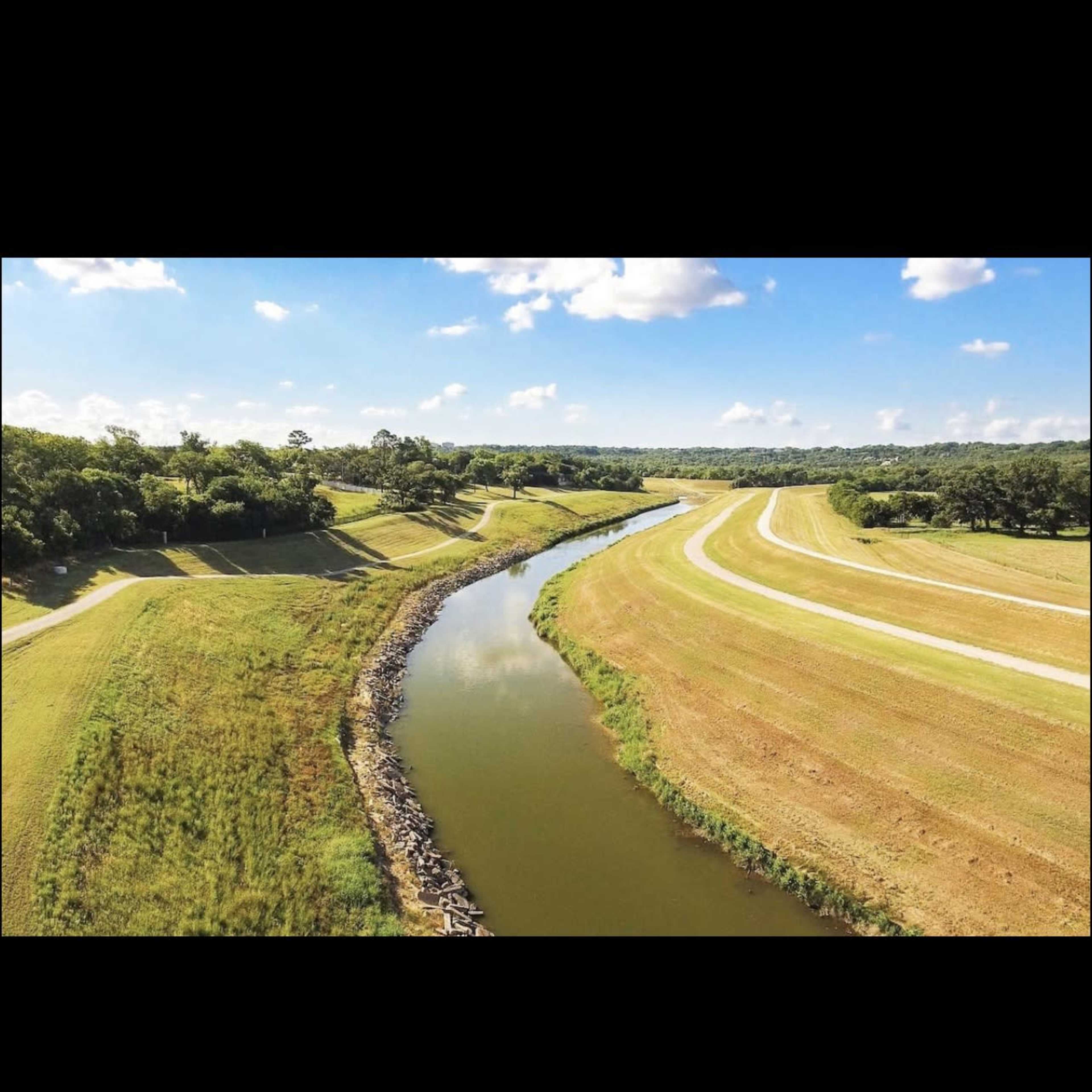 A winding river bordered by grassy fields and a pathway under a clear blue sky.