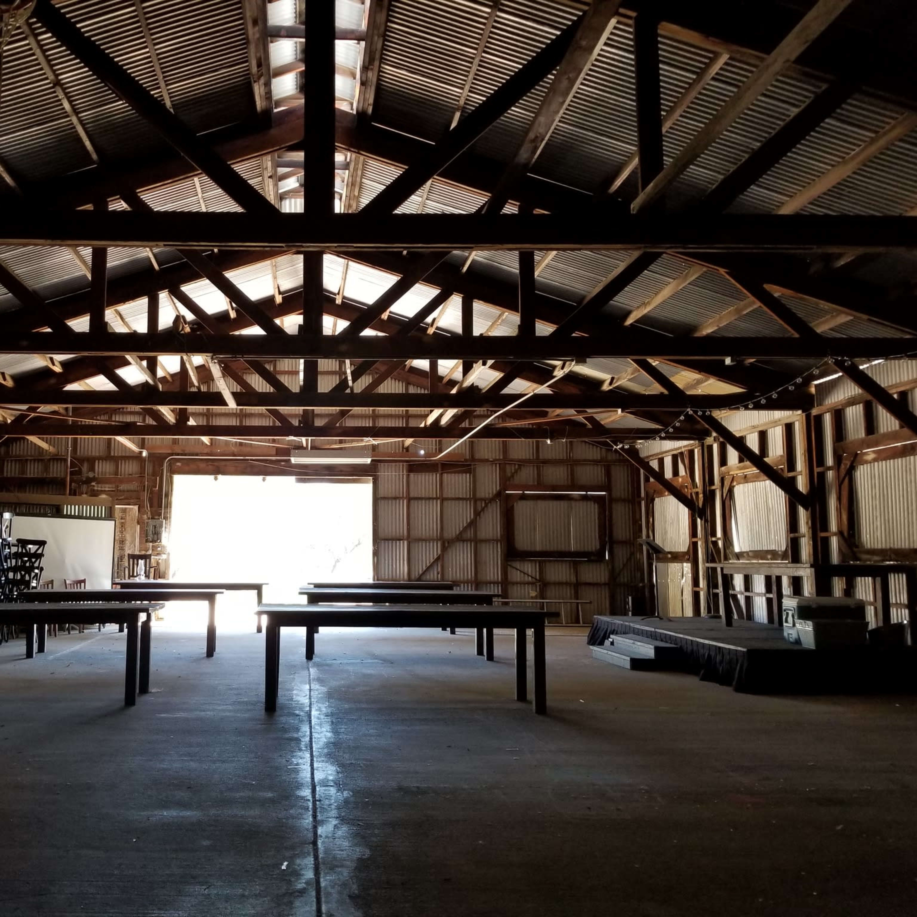 The interior of a large barn features exposed wooden beams, empty tables, and sunlight streaming in through an open door.